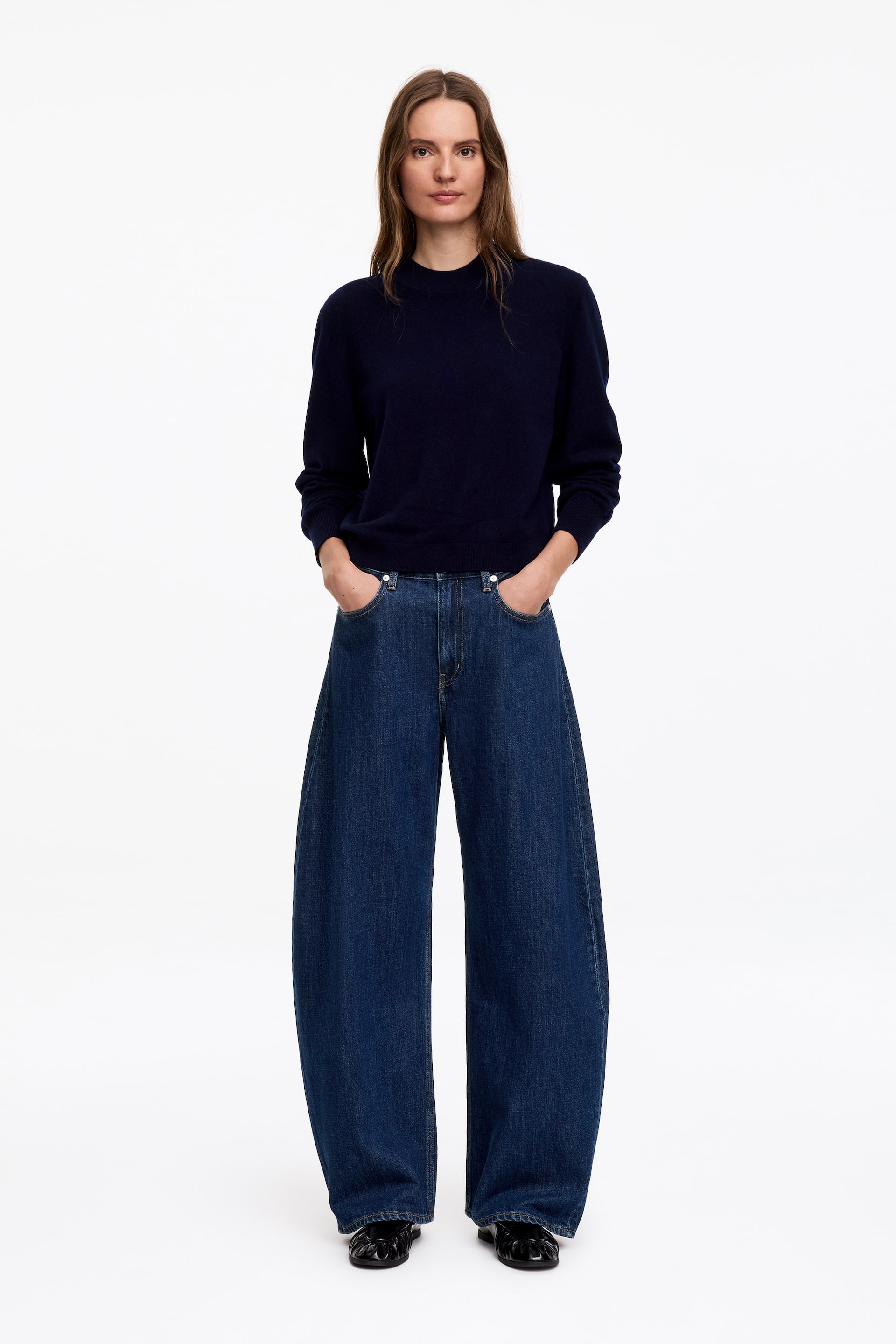 Ver imagem maior: A modelo está a usar uma camisola azul-marinho com decote redondo, combinada com jeans de perna larga em lavagem escura. A modelo coloca as mãos nos bolsos frontais dos jeans e está a usar mocassins pretos.