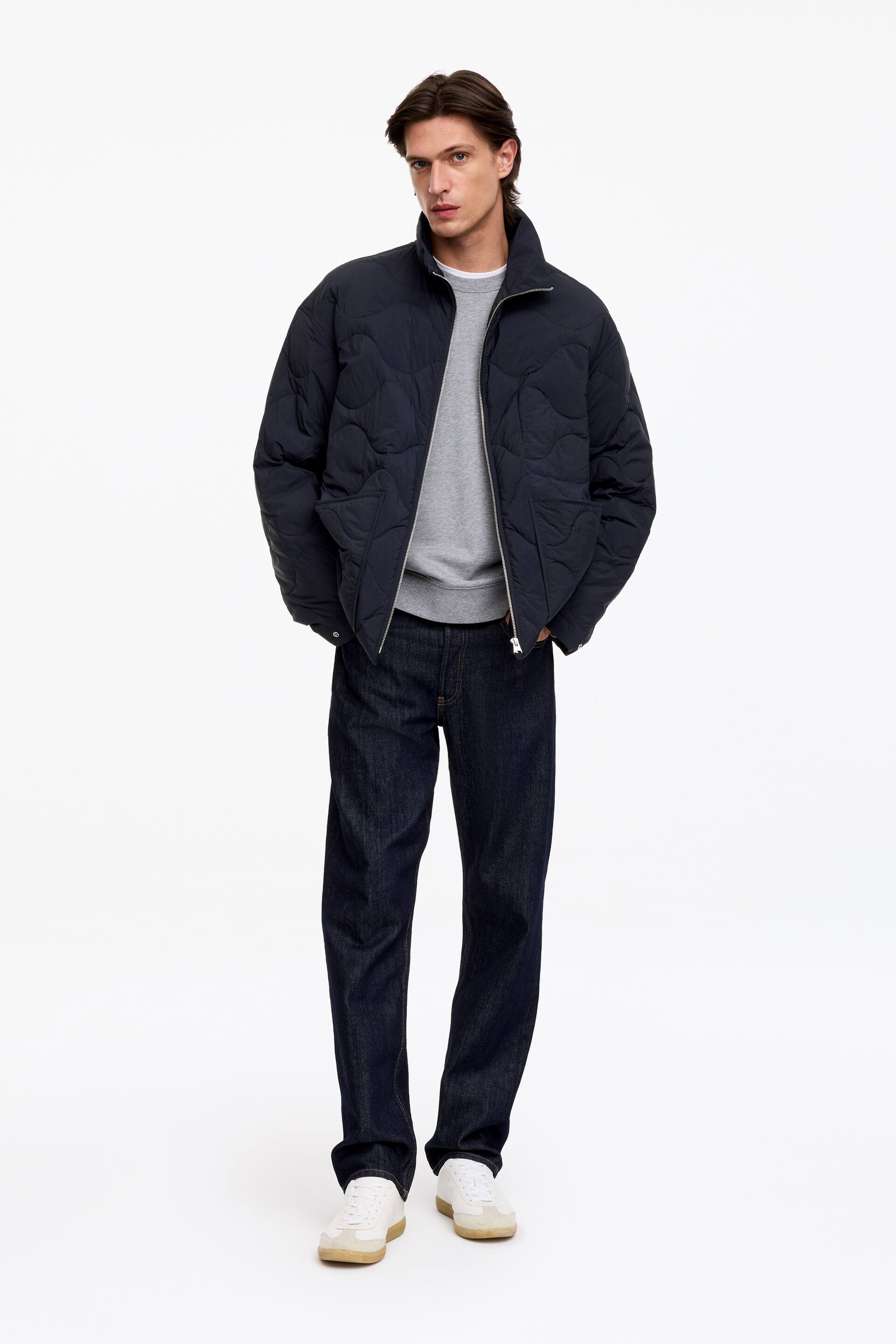 Agrandir l'image: La mannequin porte une doudoune matelassée bleu marine, présentant un motif matelassé ondulé, un col montant et une fermeture éclair frontale laissée partiellement ouverte. La veste est portée sur un sweatshirt gris et associée à un jean brut foncé.