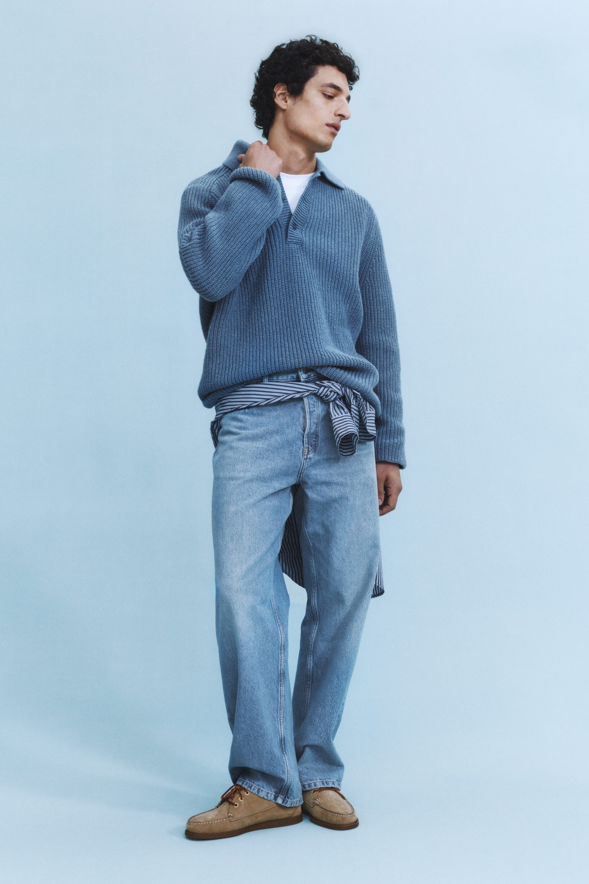 Visualizza immagine più grande: Un maglione polo blu a maglia viene presentato, caratterizzato da un colletto polo, un'apertura a V aperta e una texture a coste verticali prominente. Presenta polsini a coste e un orlo a coste, ed è stratificato sopra una t-shirt bianca con una camicia a righe annodata in vita.