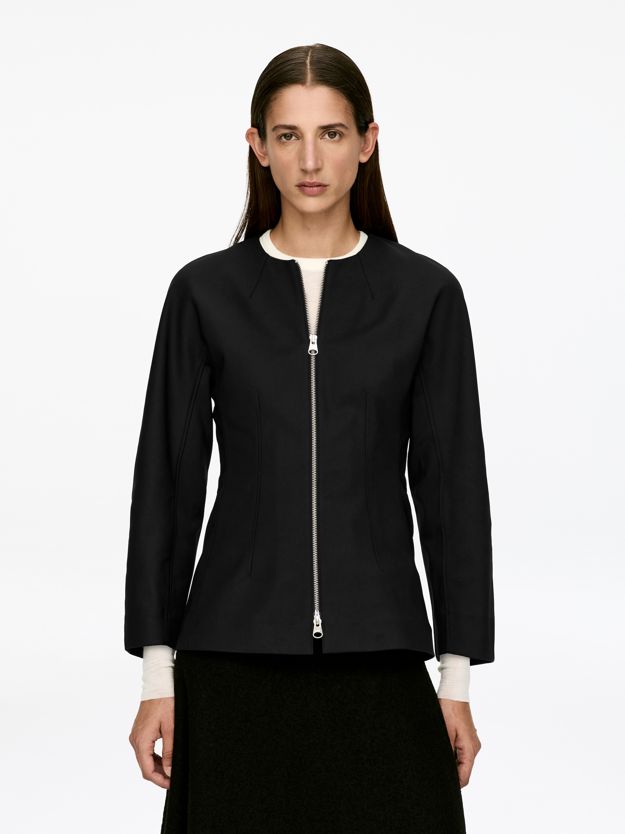 Visa större bild: En svart peplum-topp bärs av modellen. Den har en figursydd midja och en silverfärgad tvåvägsdragkedja framtill, vilken är delvis stängd. En ljusfärgad långärmad topp är synlig under, och en mörk, texturerad kjol bärs under peplumtopen.