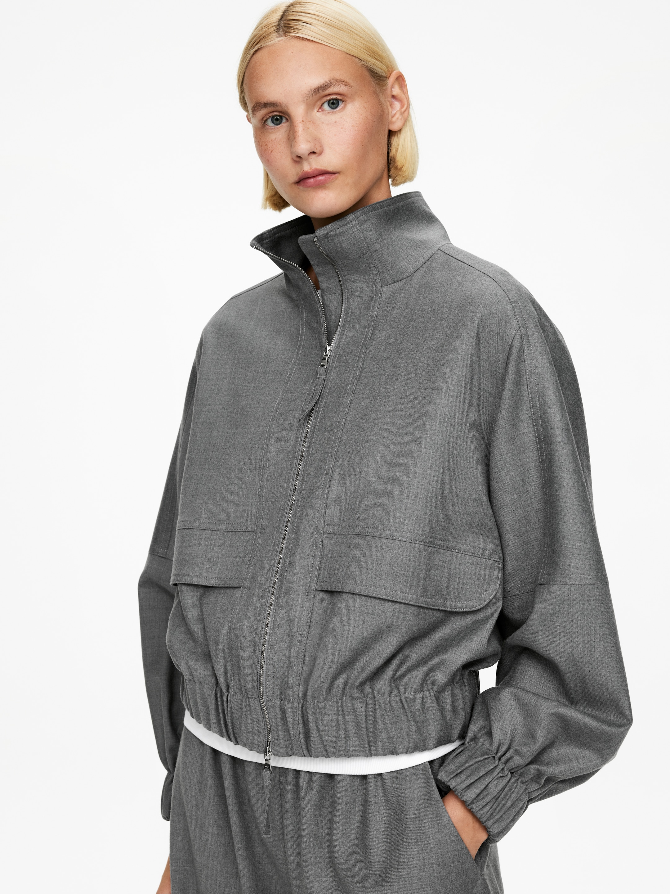 Visa större bild: En grå jacka presenteras, med en hög ståkrage, en full dragkedja framtill och två fickor med lock på bröstet. Jackan är designad med en avslappnad, kort passform, med en elastisk fåll och rynkade muddar, med en vit topp som kikar fram under.