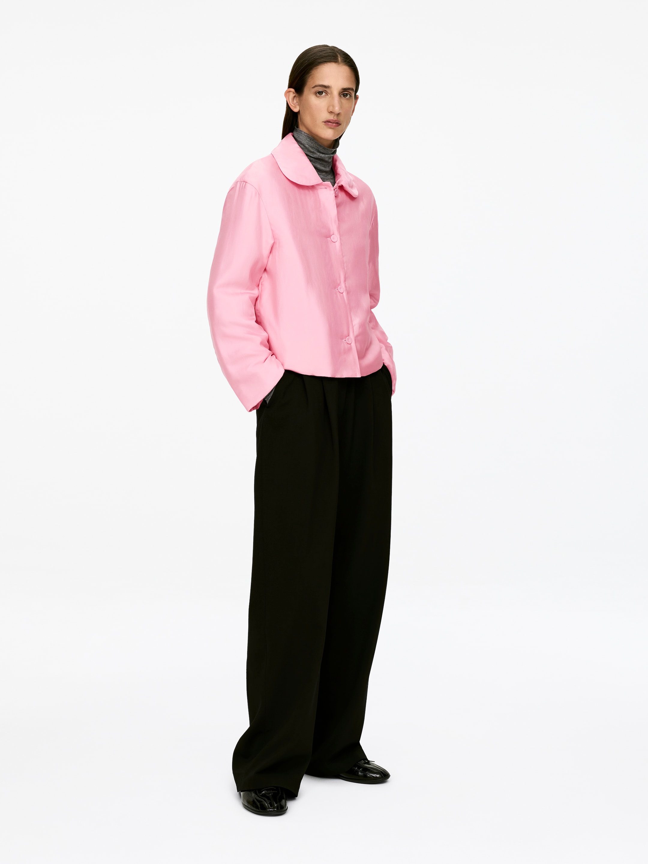 Vis større bilde: En rosa quiltet overskjorte, med en avrundet krage, stoffdekkede knapper og en vanlig passform, bæres av modellen. Den er lagd over en grå turtleneck, og modellen står med hendene i lommene.