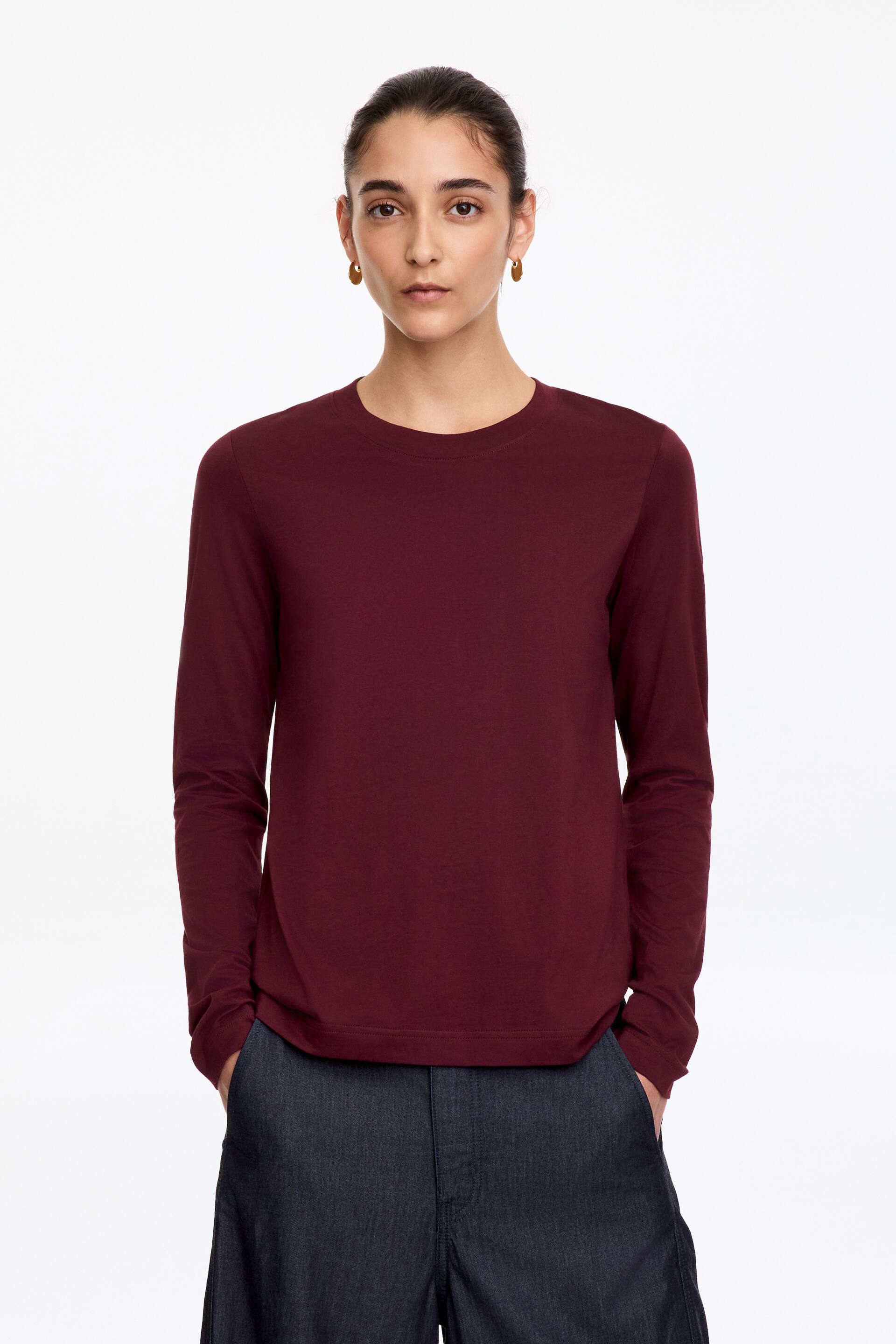 Vezi imaginea mai mare: Un tricou vișiniu cu mânecă lungă, cu guler rotund ribat și mâneci necătușate, este purtat de model, care are mâinile în buzunarele pantalonilor gri închis.
