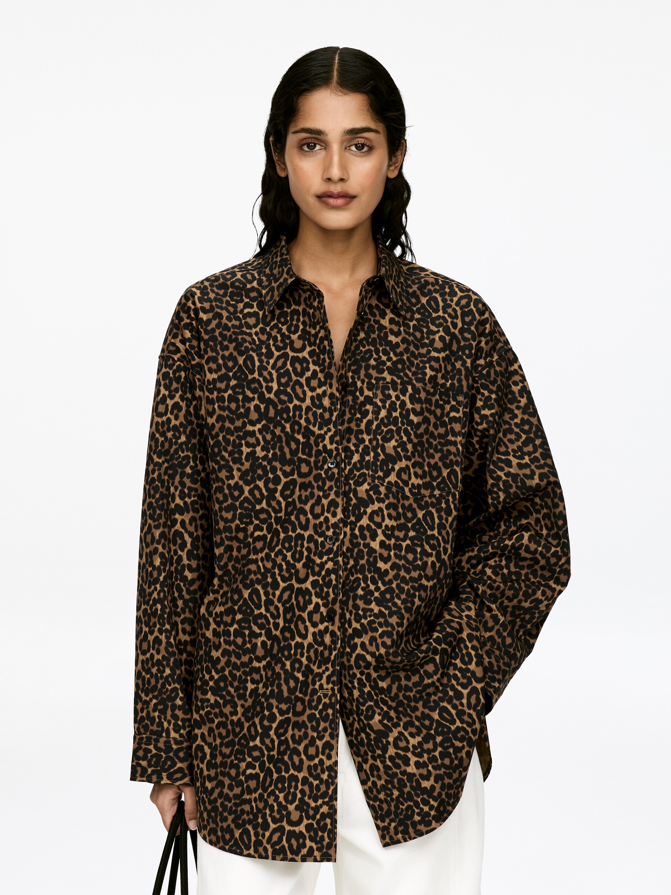 Größeres Bild ansehen: Ein übergroßes Hemd mit Leopardenmuster, spitzem Kragen und breiten, tief angesetzten Ärmeln wird von dem Model getragen. Es verfügt über eine einzelne Brusttasche, und eine der Hände des Models ruht in ihrer Tasche, während die andere eine dunkle Tasche trägt.