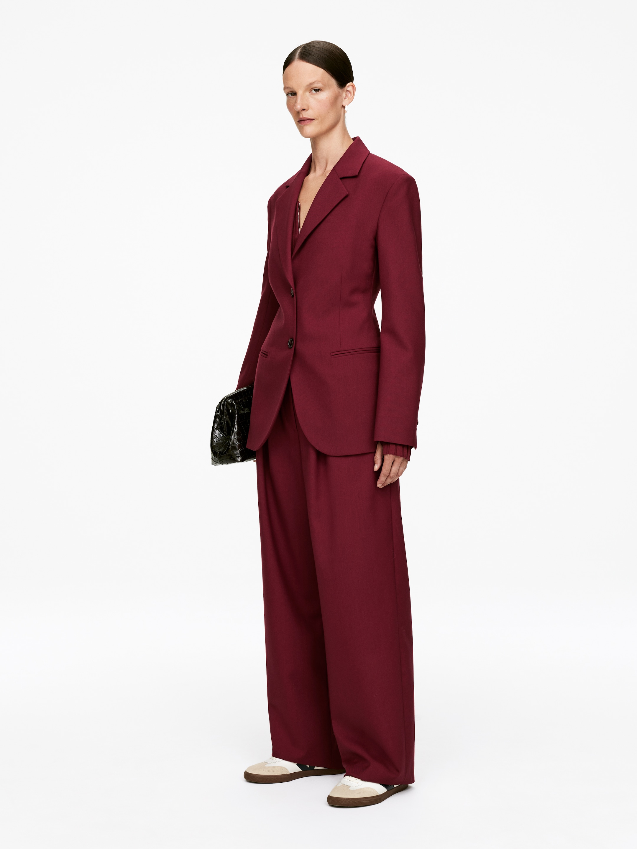 Visualizza immagine più grande: Un tailleur pantalone bordeaux è in mostra, rivolto leggermente verso destra. Presenta una giacca monopetto con revers a tacca, chiusura a un bottone e tasche a pattina, abbinata a pantaloni a gamba larga. Un top a maniche lunghe coordinato con polsini a coste è stratificato sotto la giacca.