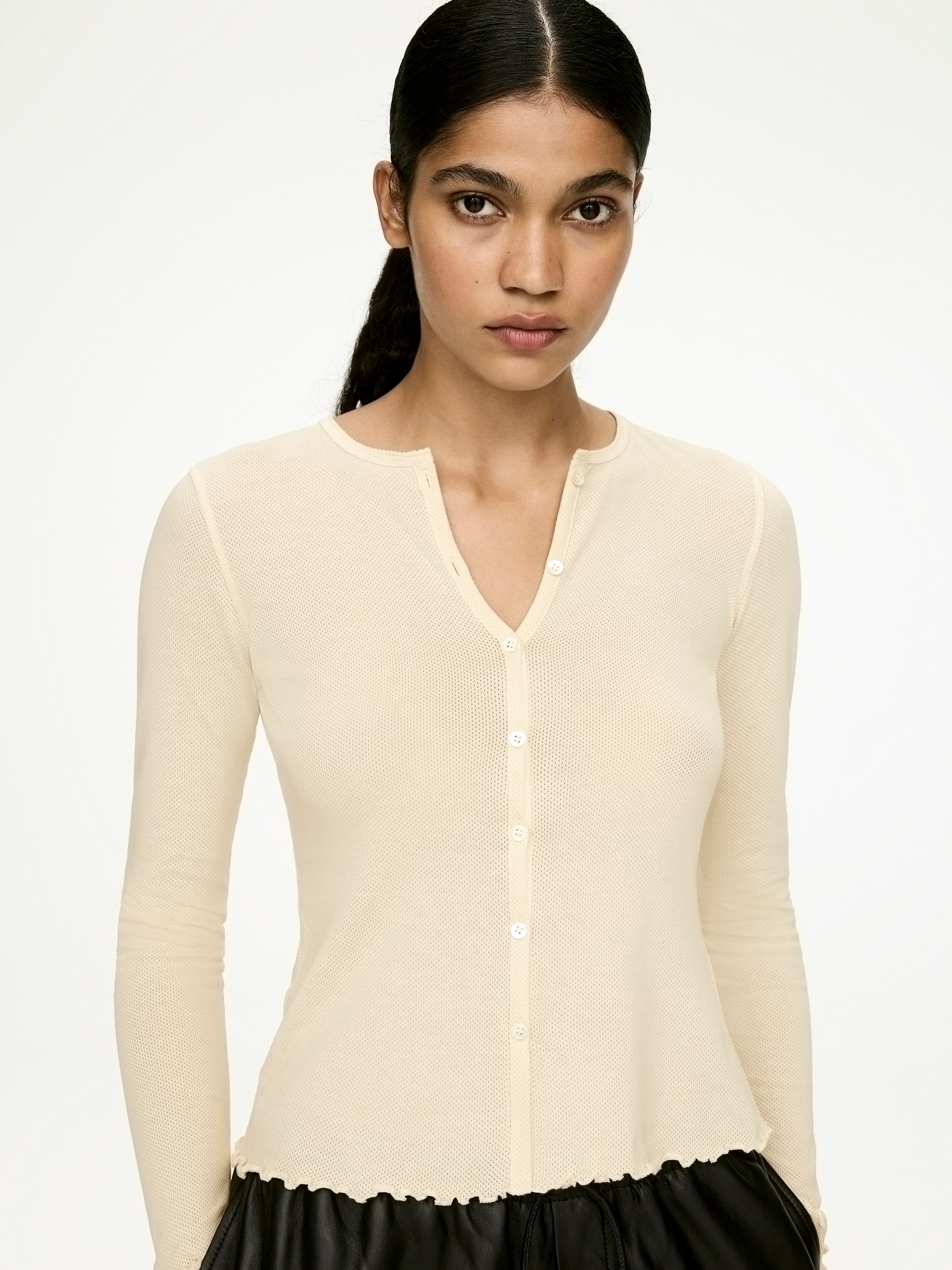 Agrandir l'image: Un haut de couleur crème en maille gaufrée, boutonné sur le devant, est orienté vers l'avant, présentant des manches longues et un décolleté en V avec une patte de boutonnage à cinq boutons. Le haut ajusté présente un ourlet volanté, visiblement superposé à un vêtement foncé.