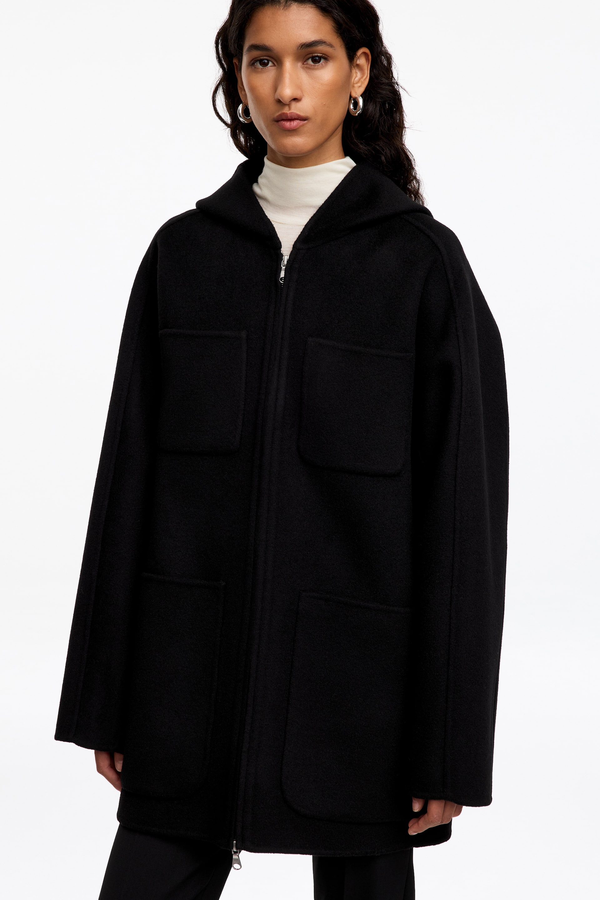 Visualizza immagine più grande: Un cappotto nero in misto lana con cappuccio, caratterizzato da una silhouette rilassata, chiusura frontale con zip e ampie tasche applicate, indossato dalla modella. Il cappotto è abbinato a un maglione bianco a collo alto.