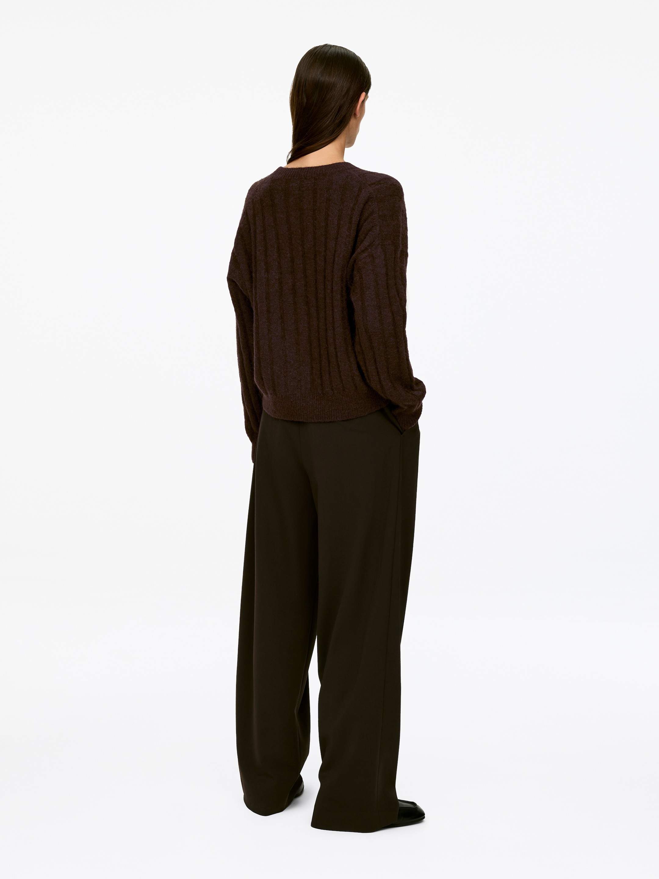 Grotere afbeelding bekijken: Een donkerbruine wijdgeribde trui met lange mouwen en een ontspannen pasvorm wordt gedragen door het model. De trui heeft verticale ribbels en is gestyled over een donkere broek met wijde pijpen.