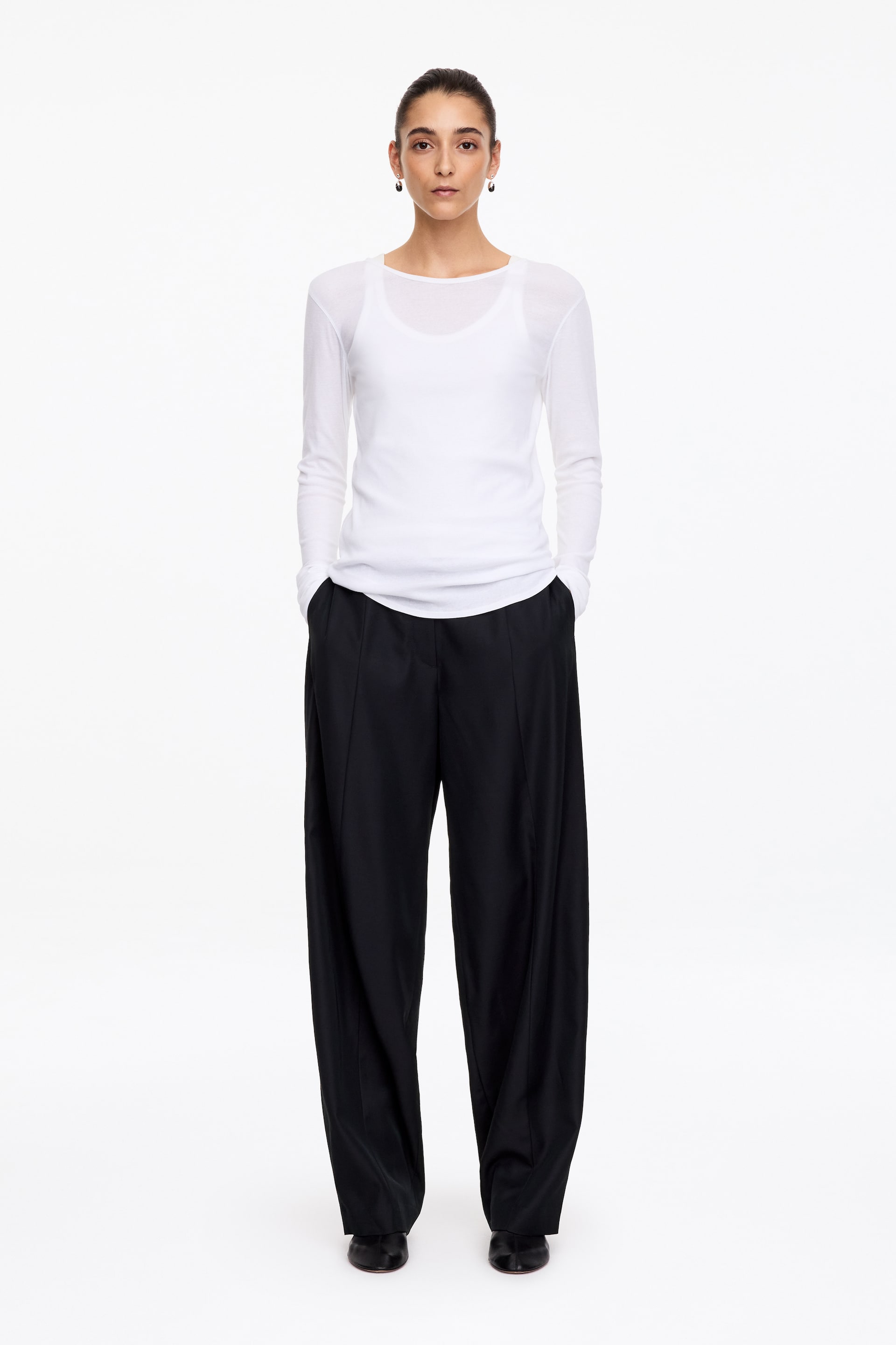 Größeres Bild anzeigen: Schwarze Barrel-Leg-Hose mit entspannter, skulpturierter Silhouette und vertikalen Bügelfalten vorn wird vom Model getragen. Die Hose ist mit einem weißen Langarm-Top kombiniert, während das Model steht und die Hände in die Seitentaschen steckt.