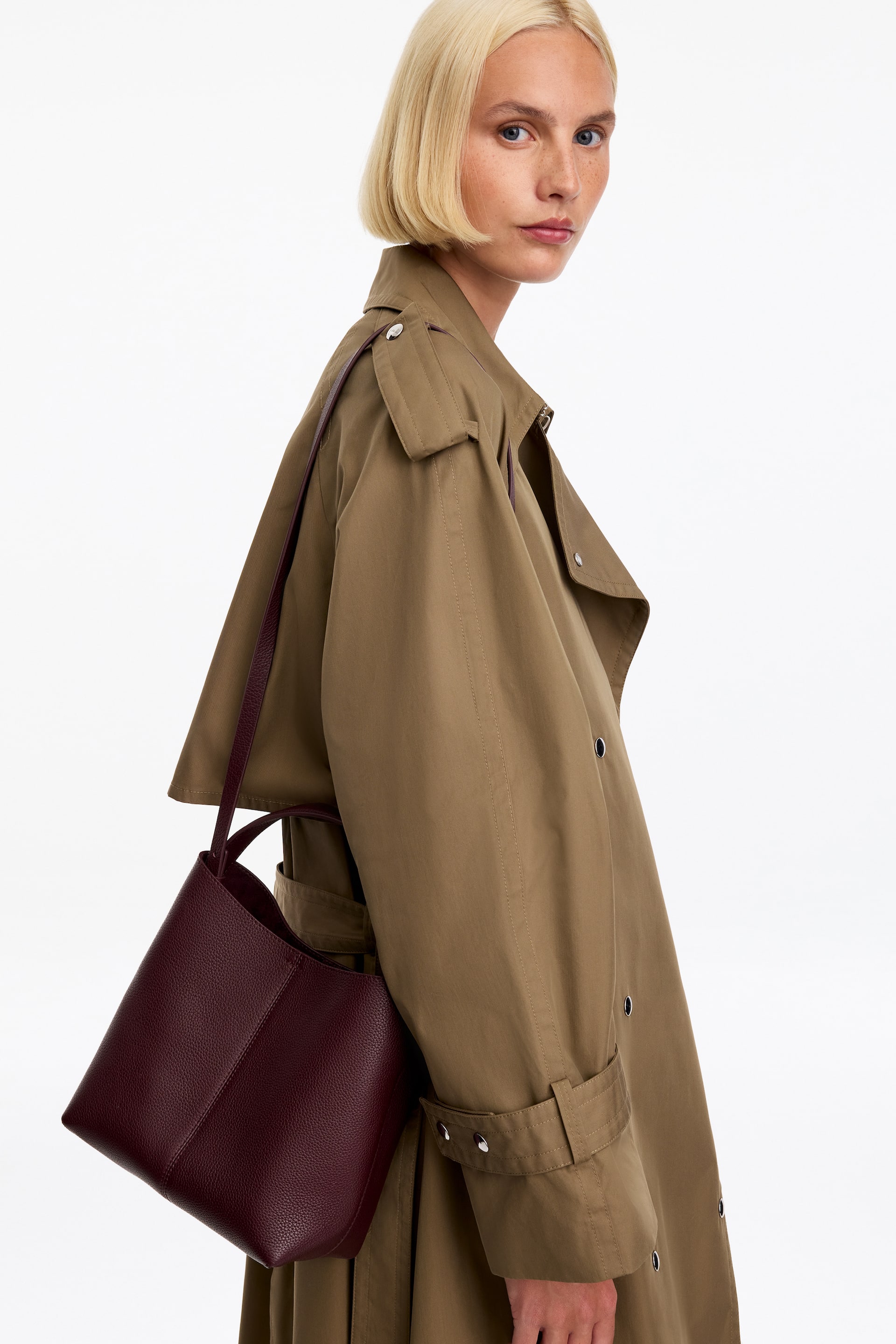 Agrandir l'image: Un petit sac seau en cuir bordeaux à la finition texturée et doté d'une fine bandoulière est porté à l'épaule par le mannequin, qui porte un trench-coat kaki. Le sac présente une silhouette souplement structurée avec des surpiqûres visibles.