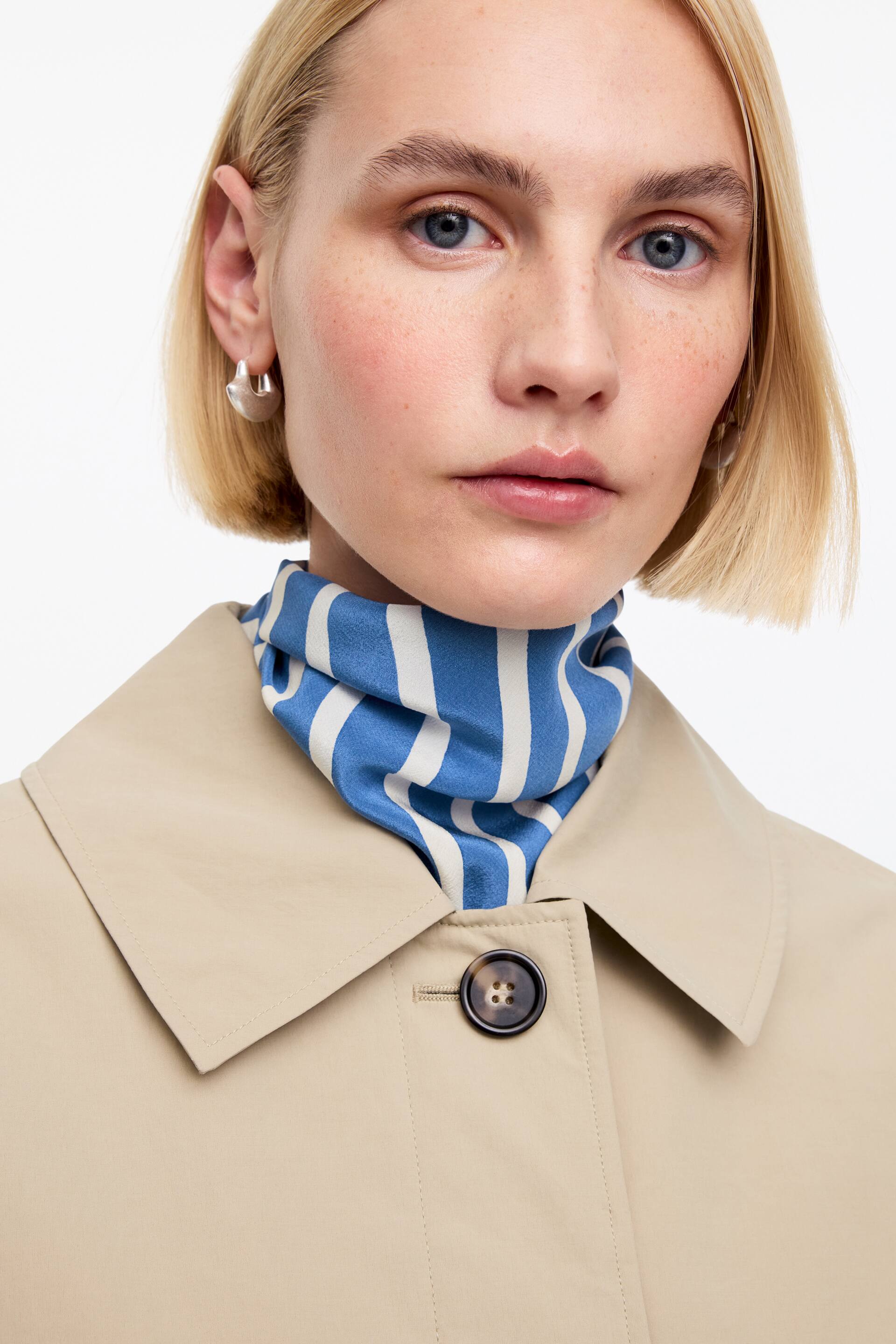 Ver imagen más grande: Una chaqueta beige presenta un clásico cuello en punta y un cierre de botones oscuros moteados en la parte delantera. Una bufanda de rayas azules y blancas está colocada alrededor del cuello, visible debajo del cuello de la chaqueta.