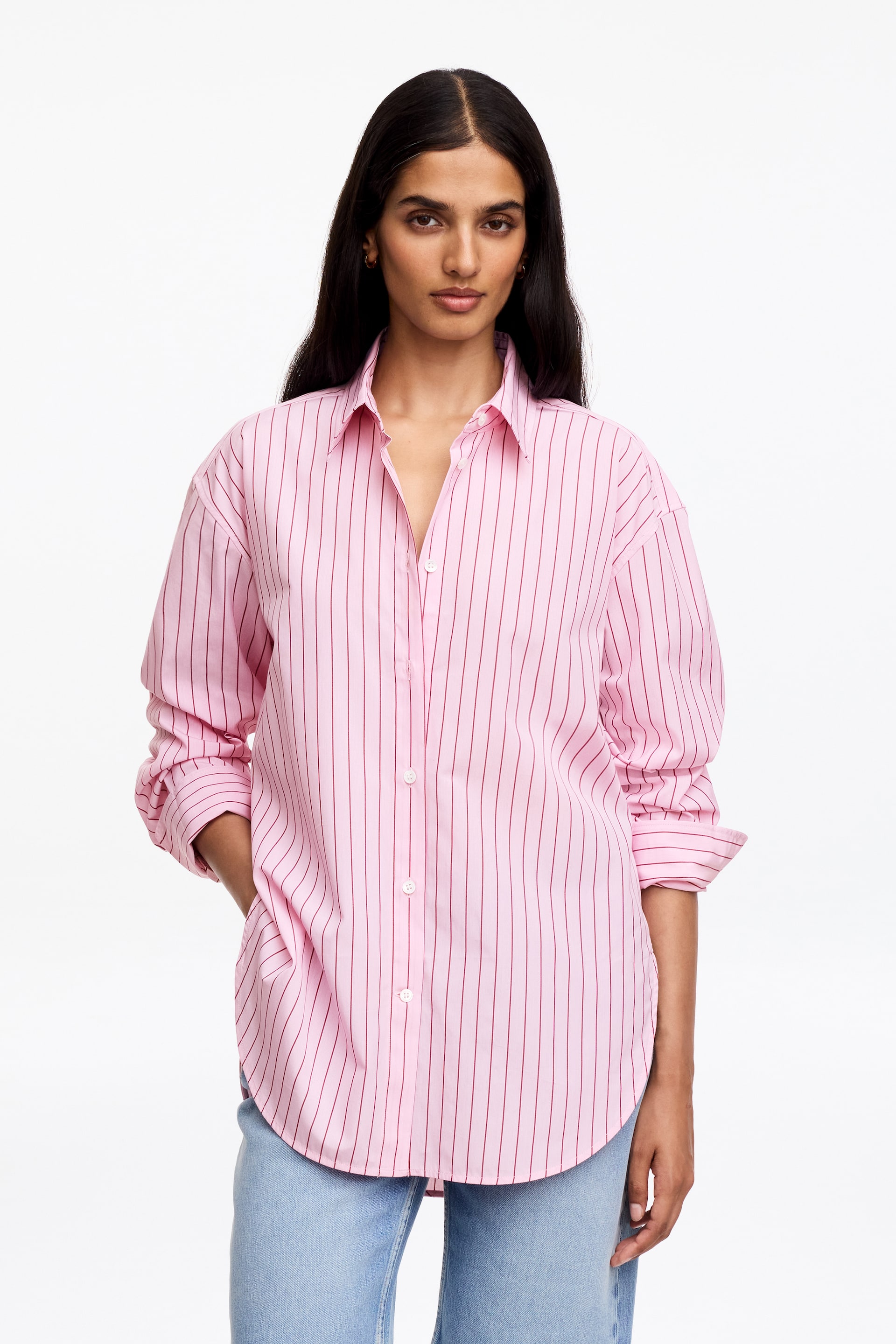 View larger image: The pink button-up shirt is facing forward, featuring thin vertical dark red stripes. It is designed with a pointed collar, a full button placket with white buttons, and long sleeves that are rolled up, presenting an oversized fit and a rounded hem.