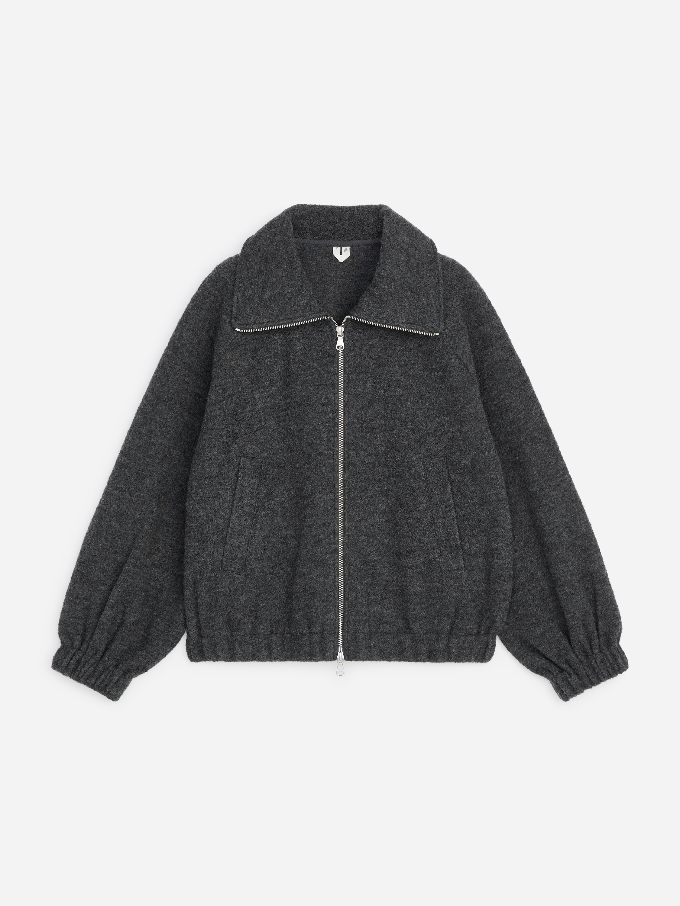 En mÃ¶rkgrÃ¥ texturerad ulljacka med silverfÃ¤rgad dragkedja framtill, bred krage samt resÃ¥rsammandragna muddar och nederkant bÃ¤rs av modellen. Modellen stÃ¥r med hÃ¤nderna vilande i sidofickorna och stylar jackan Ã¶ver en grÃ¥ ribbstickad polotrÃ¶ja och svarta byxor.