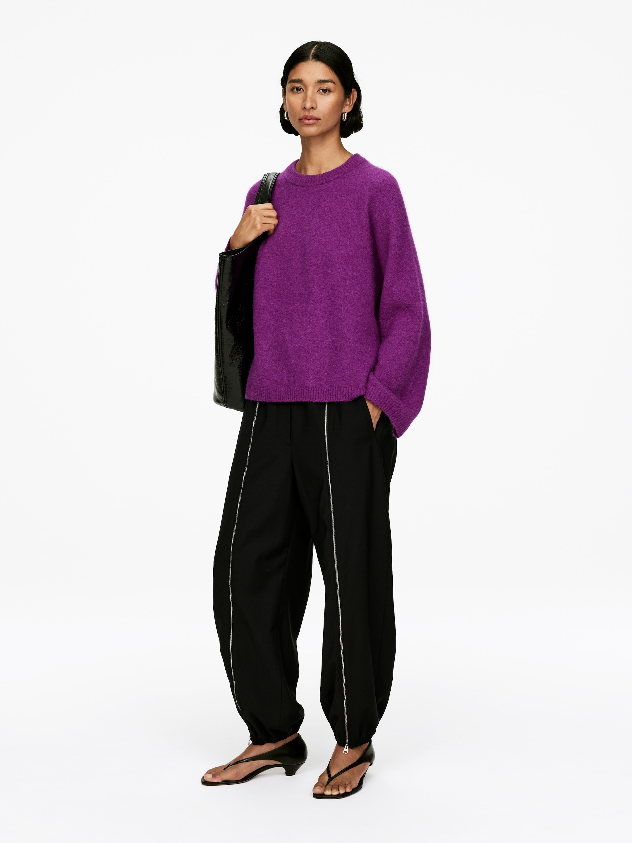 Agrandir l'image: Un pull à col rond violet vif est présenté, avec une texture duveteuse, une coupe décontractée et des manches longues volumineuses avec des poignets côtelés. Un pantalon noir à jambes larges est également porté, mettant en valeur des fermetures éclair argentées proéminentes qui descendent verticalement sur le devant de chaque jambe jusqu'aux chevilles élastiquées et froncées.