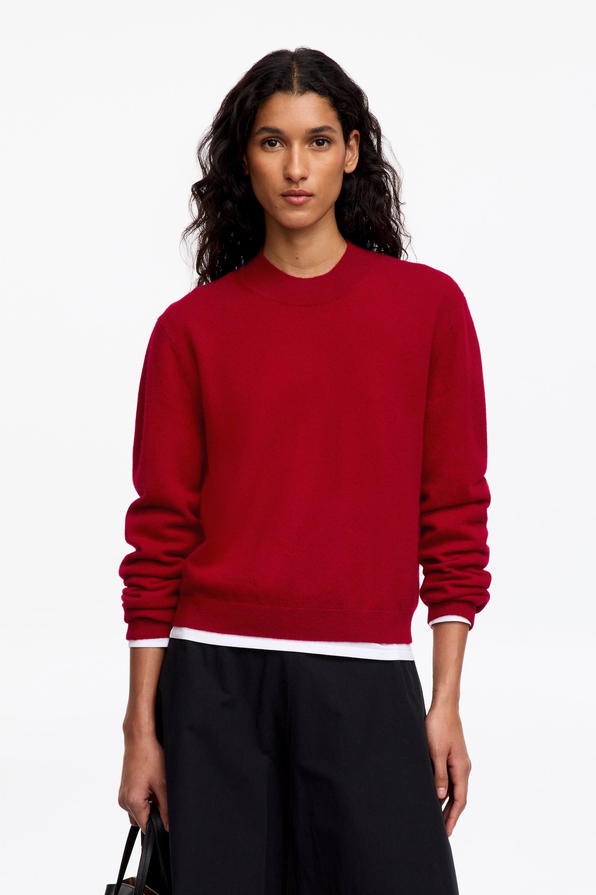 Agrandir l'image: Le mannequin porte un pull en maille rouge à col rond. Il présente des manches longues et est superposé à un T-shirt blanc, visible au niveau de l’ourlet et des poignets.