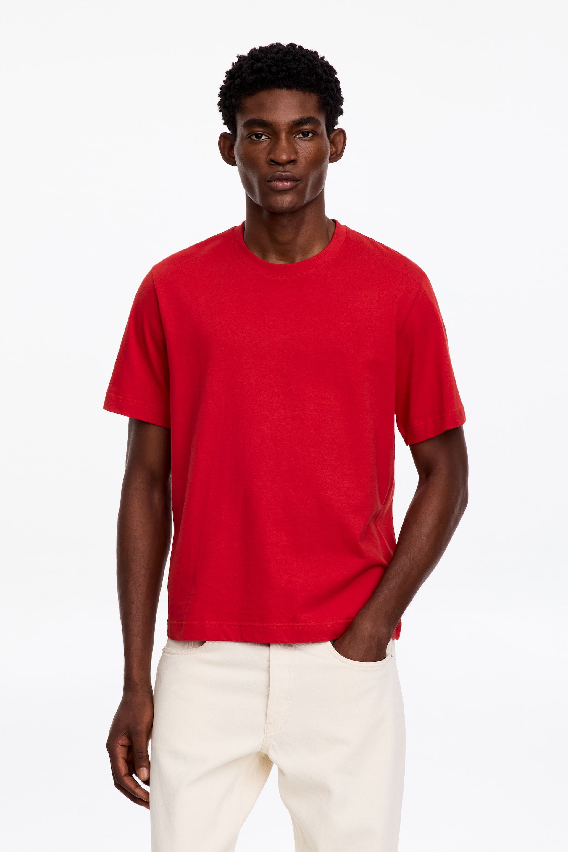 Vezi imaginea mai mare: Un tricou roșu cu mânecă scurtă și guler rotund este purtat de model, care îl asortează cu pantaloni de culoare crem.