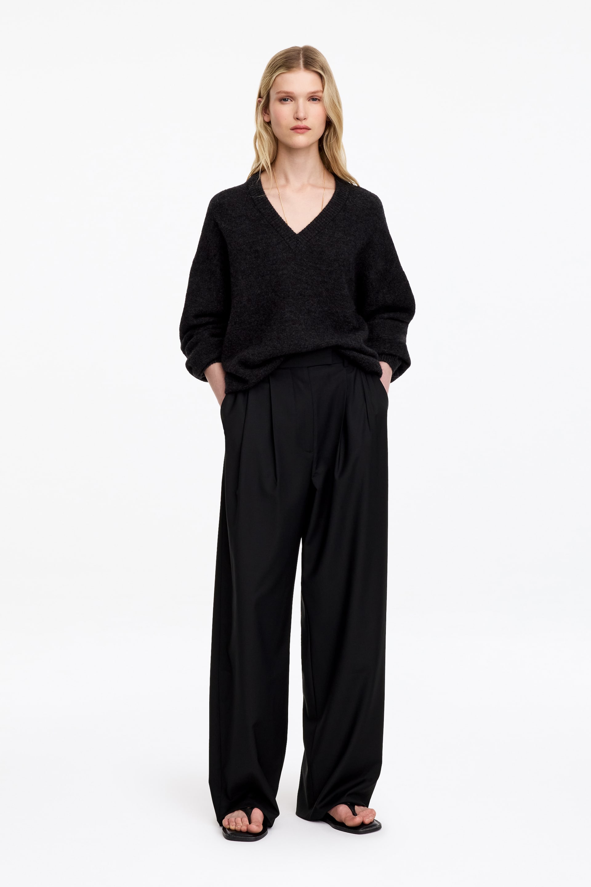 Se større billede: Sorte højtaljede bukser med vide ben og læg foran bæres af modellen, som står med hænderne hvilende i sidelommerne. Bukserne er stylet med en sort V-hals sweater, der er stukket ned foran i linningen, og kombineret med sorte sandaler.
