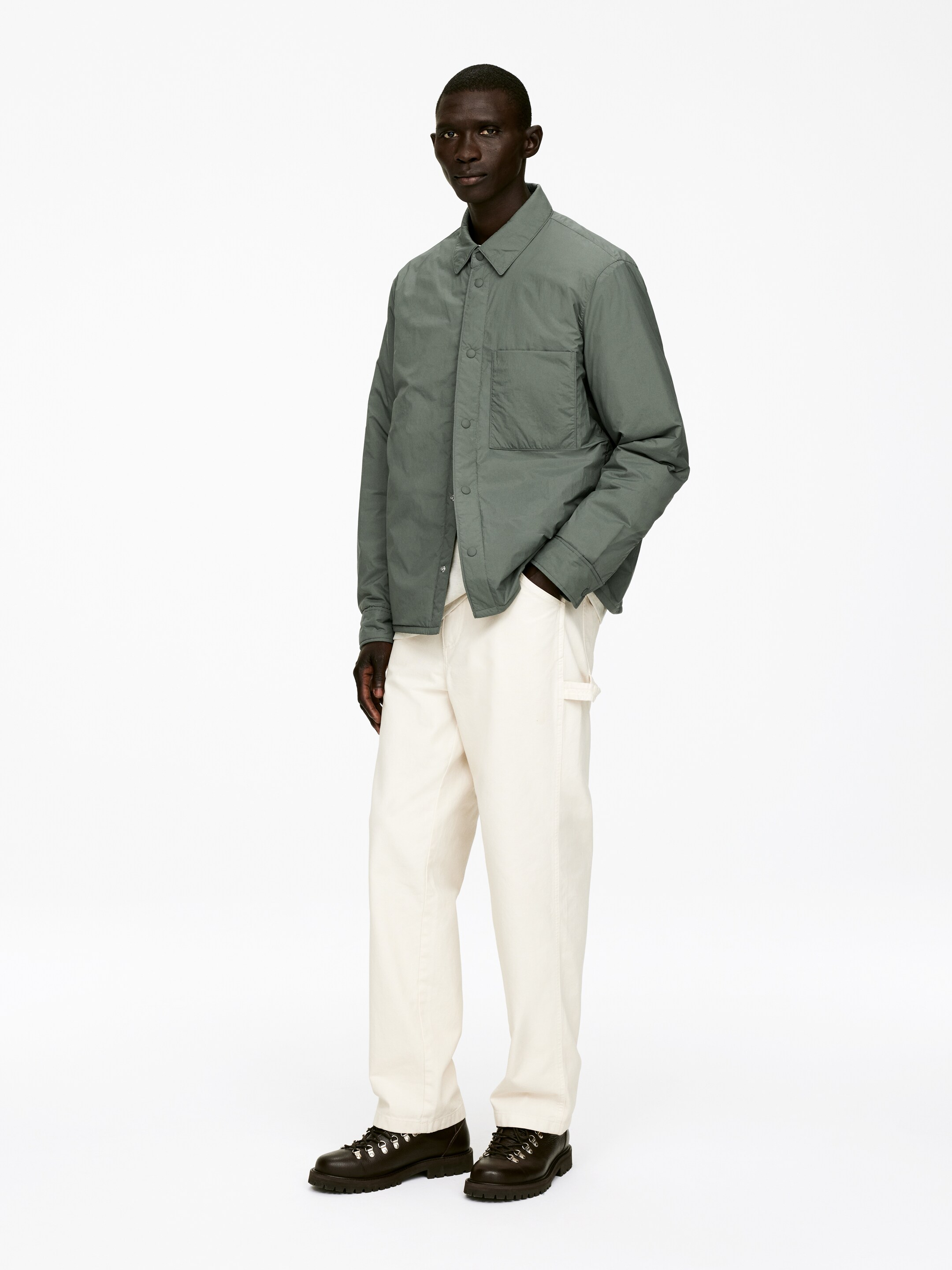 Ver imagen más grande: Una chaqueta de camisa acolchada de color verde apagado se presenta desde una vista frontal ligeramente angulada, con un cuello de punta, un cierre de botones en la parte delantera y un único bolsillo de parche en el pecho. Se lleva sobre unos pantalones de carpintero de color blanco roto, que muestran un lazo para martillo y un bolsillo lateral para herramientas, y botas de cordones de color marrón oscuro que presentan ojales en forma de D y suelas gruesas con tacos.