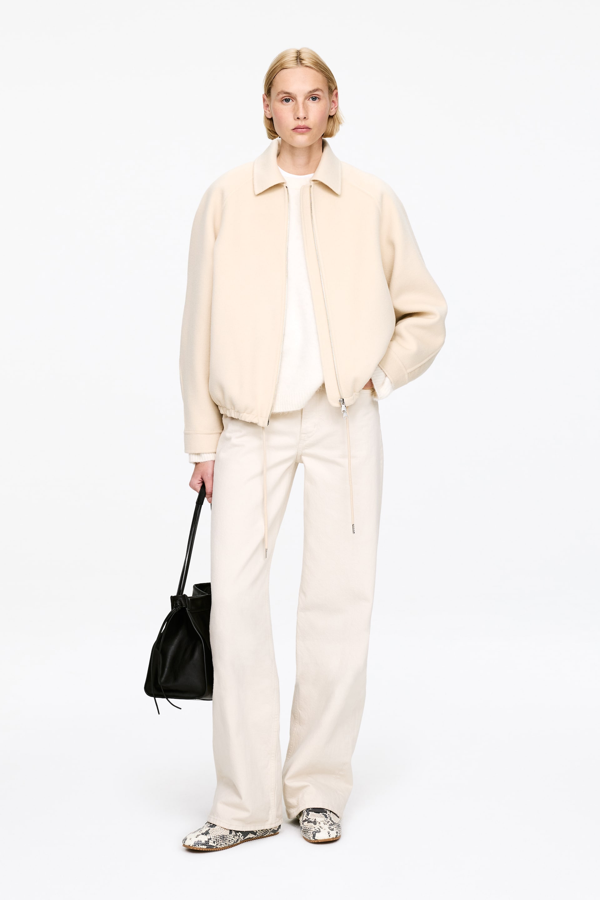 Ver imagem maior: Um blusão creme está a ser apresentado, de frente, com fecho completo à frente, gola pontiaguda, bolsos laterais e bainha elástica. Um camisola creme é visível por baixo.