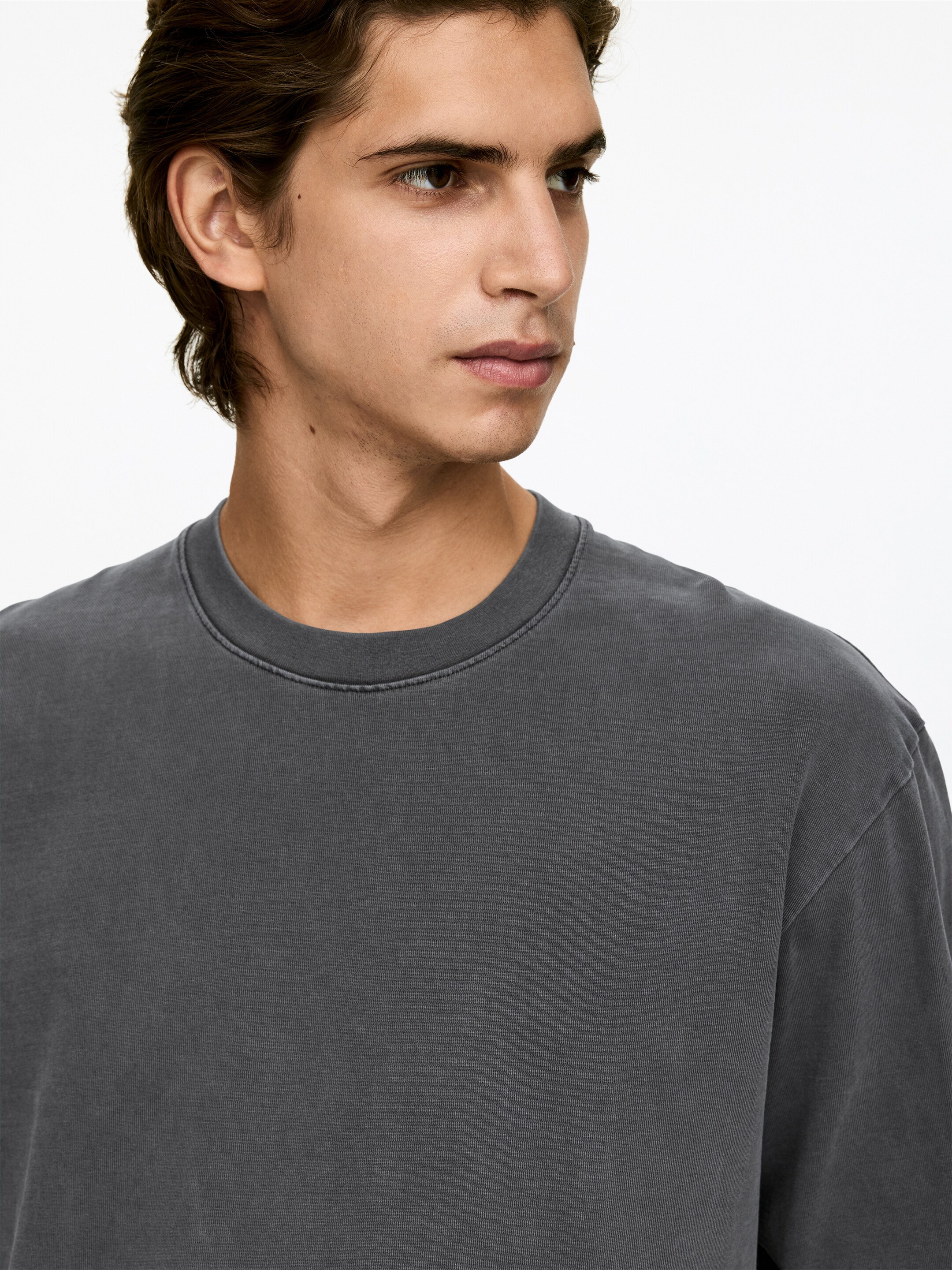 View larger image: A dark gray crew-neck t-shirt is being worn, presenting a washed appearance to its fabric. The round neckline is featuring a double-stitched collar band, and a relaxed fit is visible across the shoulders.