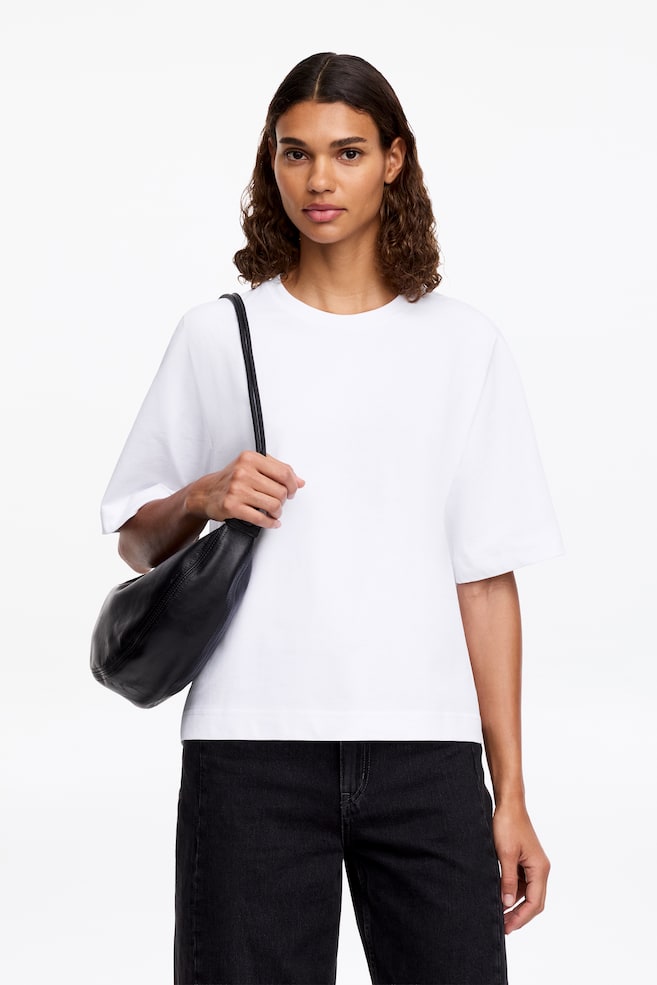 Una camiseta blanca se muestra desde el frente, con un cuello redondo y mangas cortas y anchas. Su corte relajado cae justo por encima de las caderas. - 1