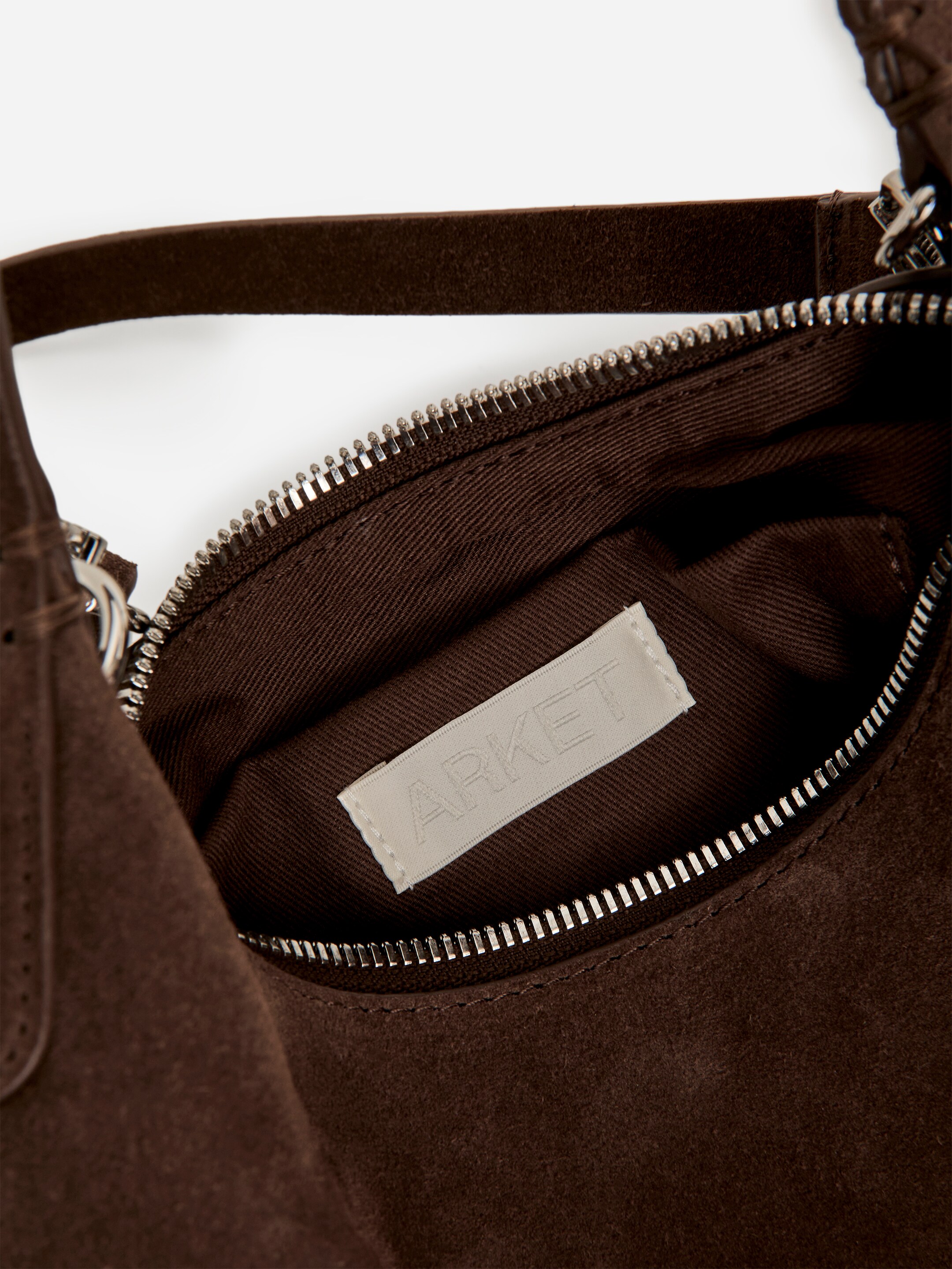 Größeres Bild ansehen: Eine dunkelbraune, quadratische Tasche aus Wildleder wird von OBEN betrachtet, sie ist offen und offenbart ihr dunkelbraunes Innenfutter aus Stoff. Ein Reißverschluss in Silberoptik sichert die obere Öffnung, mit einem hellen Markenetikett, das innen sichtbar ist, und einem dunkelbraunen Wildledertragegriff, der von der Seite ausgeht.