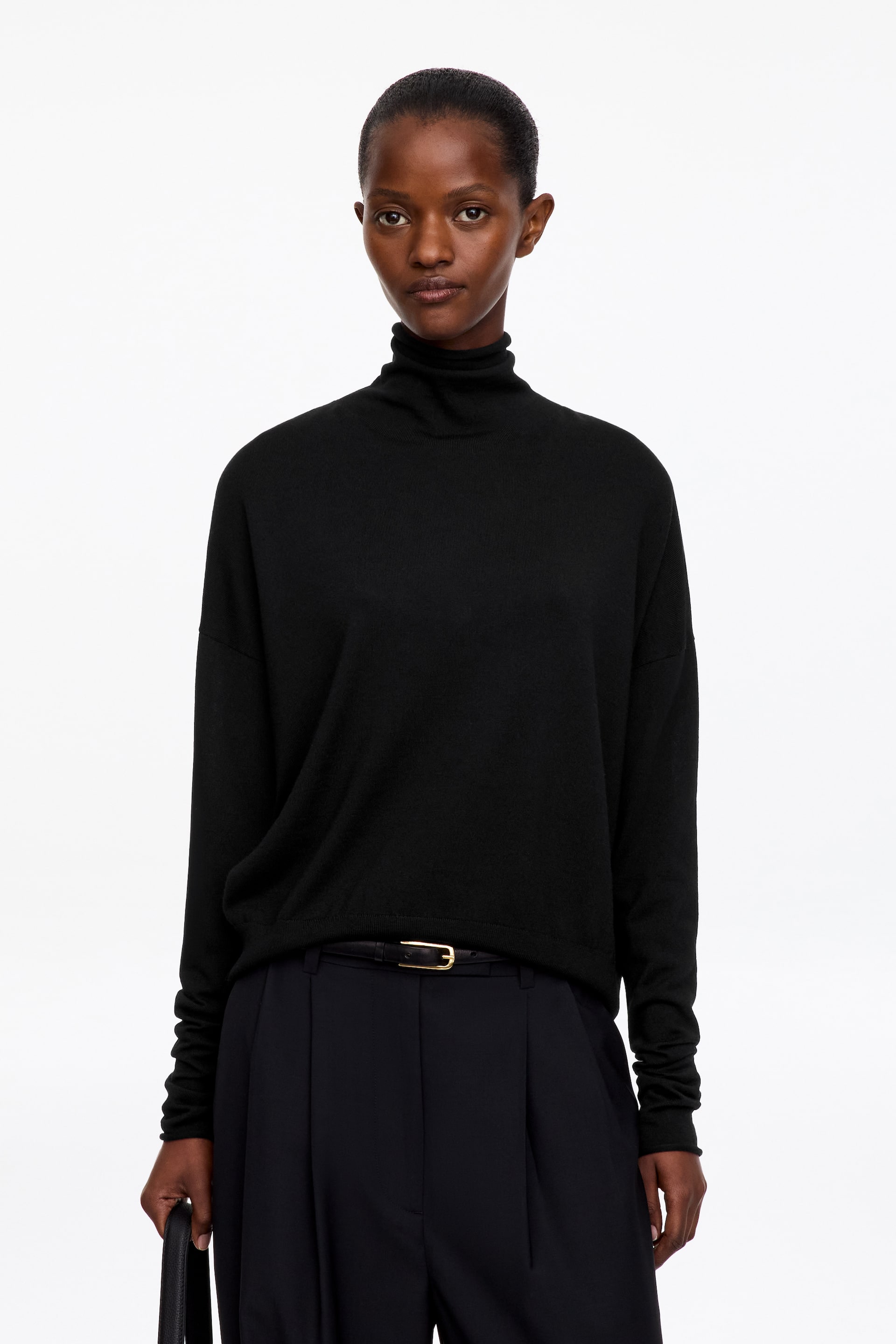 Visualizza immagine più grande: La modella indossa un maglione nero a collo alto in maglia fine, dal taglio rilassato con spalle scese e maniche allungate che si raccolgono ai polsi. Il maglione è abbinato a pantaloni neri plissettati e a una sottile cintura con fibbia dorata.