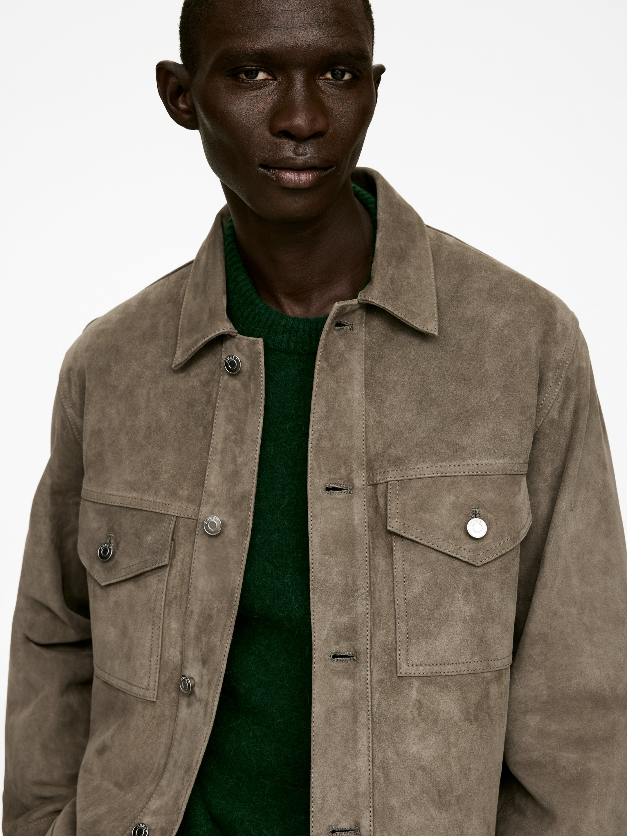 Agrandir l'image: Une veste-chemise en daim taupe est portée par-dessus un pull à col rond vert foncé. La veste présente un col classique à pointes, une fermeture à boutons sur le devant et deux poches poitrine avec des boutons-pression visibles sur les rabats.