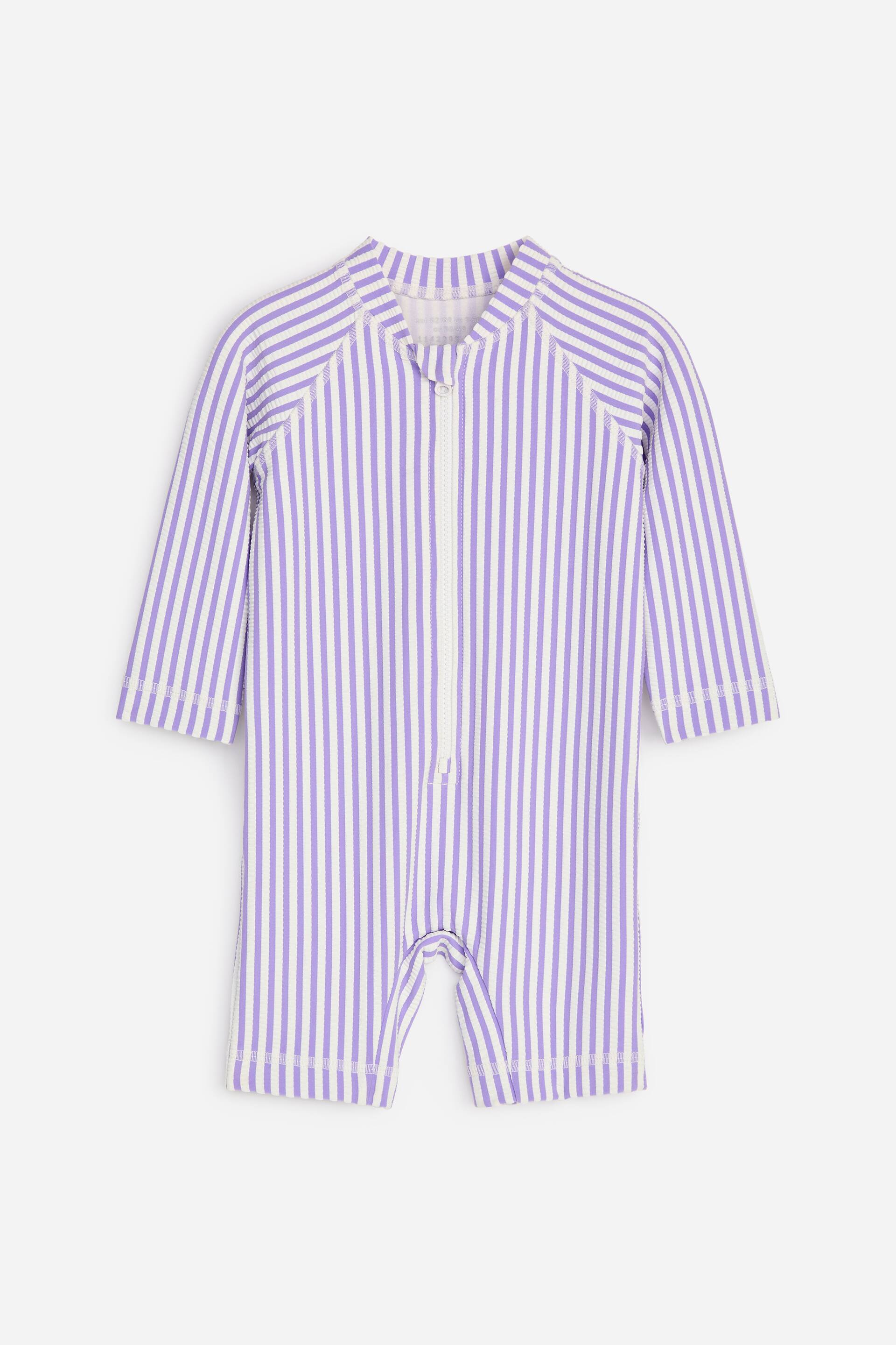 Agrandir l'image: Une combinaison en seersucker à manches longues est présentée de face, ornée de rayures verticales violettes et blanches. Elle est conçue avec un col montant, une fermeture éclair blanche qui s'étend au centre, et des ouvertures de jambes courtes.
