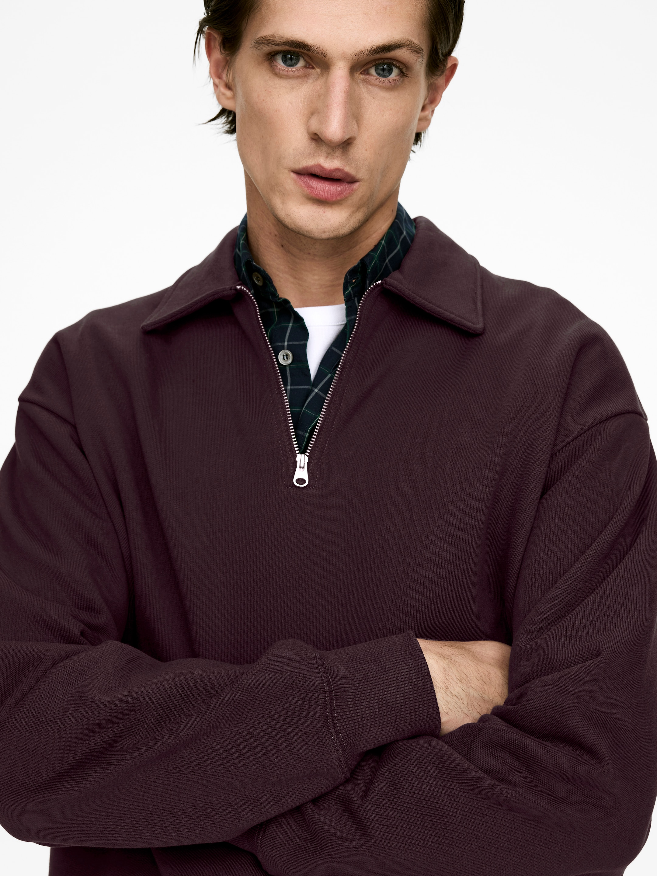 Agrandir l'image: Un pull-over à demi-fermeture éclair de couleur violet foncé est porté, avec un col évasé, une fermeture éclair partiellement ouverte de couleur argentée, et des poignets côtelés sur ses manches longues. Il est superposé à un t-shirt blanc et une chemise à carreaux foncés, offrant une coupe décontractée.
