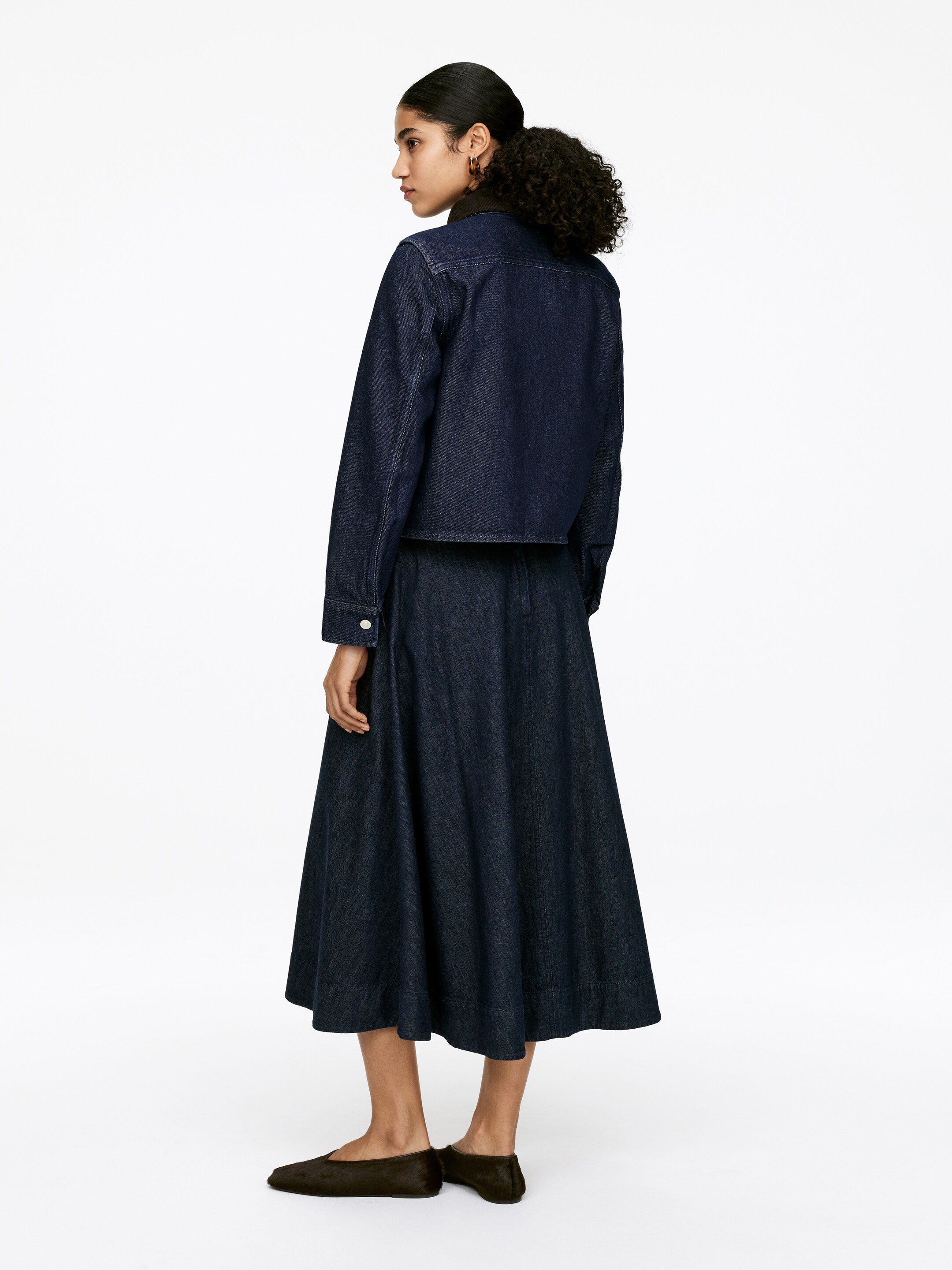 Ver imagen más grande: Una sobrecamisa de mezclilla índigo oscuro, con un cuello de pana y un ajuste regular, está siendo llevada por la modelo. La sobrecamisa termina en la cintura y se está combinando con una falda midi de mezclilla oscura a juego.