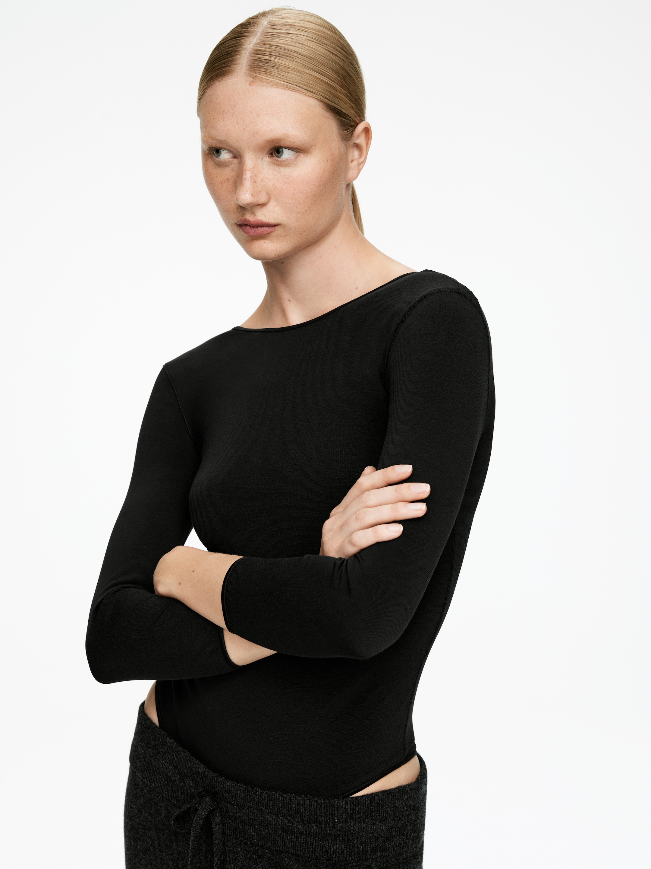 Visa större bild: En svart, figurnära bodysuit, tillverkad av ett stretchigt tyg, bärs av modellen. Den har långa ärmar och en bred båtringning. Modellen korsar armarna, och mörka byxor är lagda över bodysuitens nedre del.