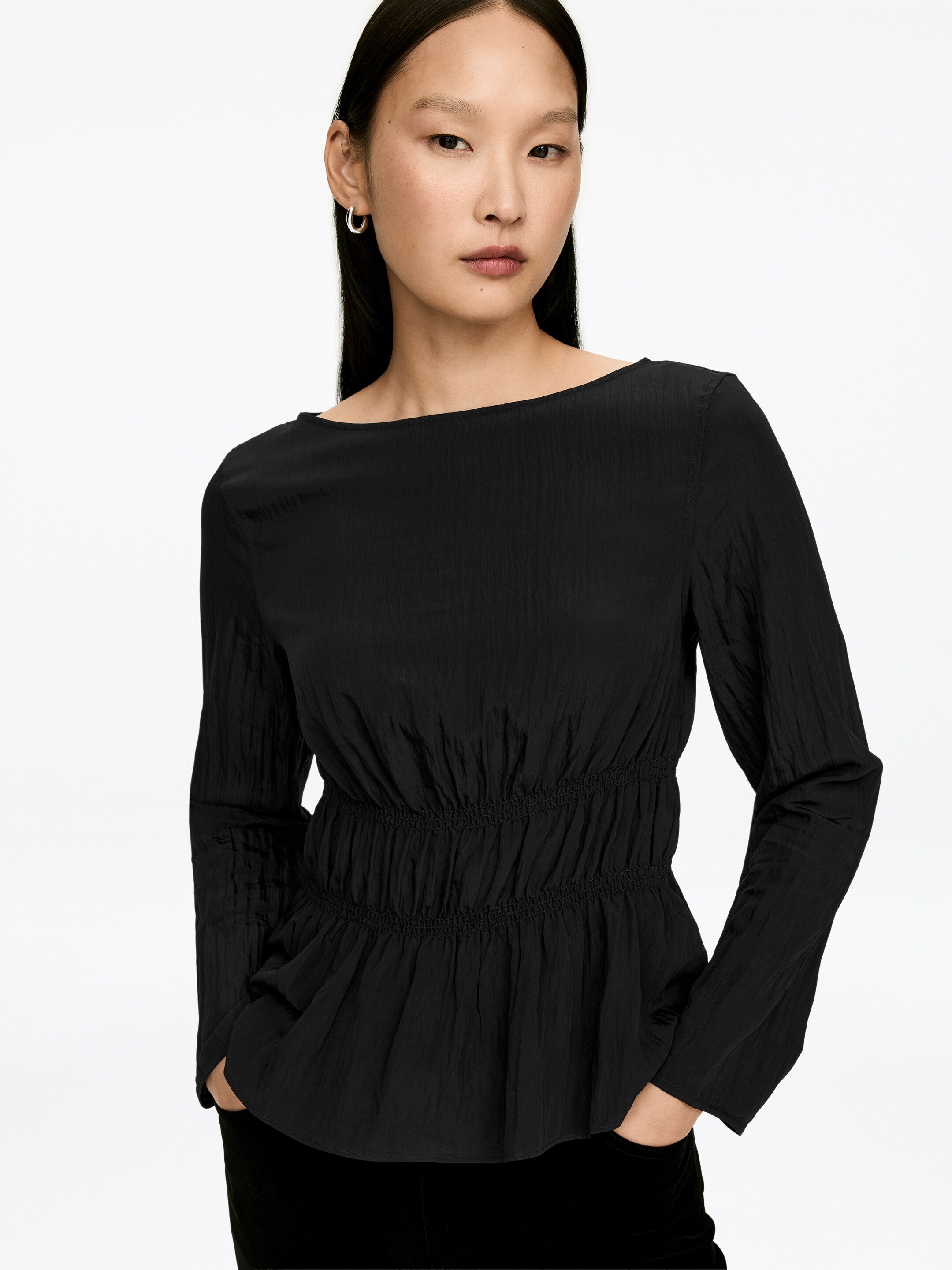 View larger image: A black textured top is featuring a wide boat neckline and long sleeves. It is showcasing a crinkled fabric with a wide shirred elasticized waist, creating a gathered peplum hem.