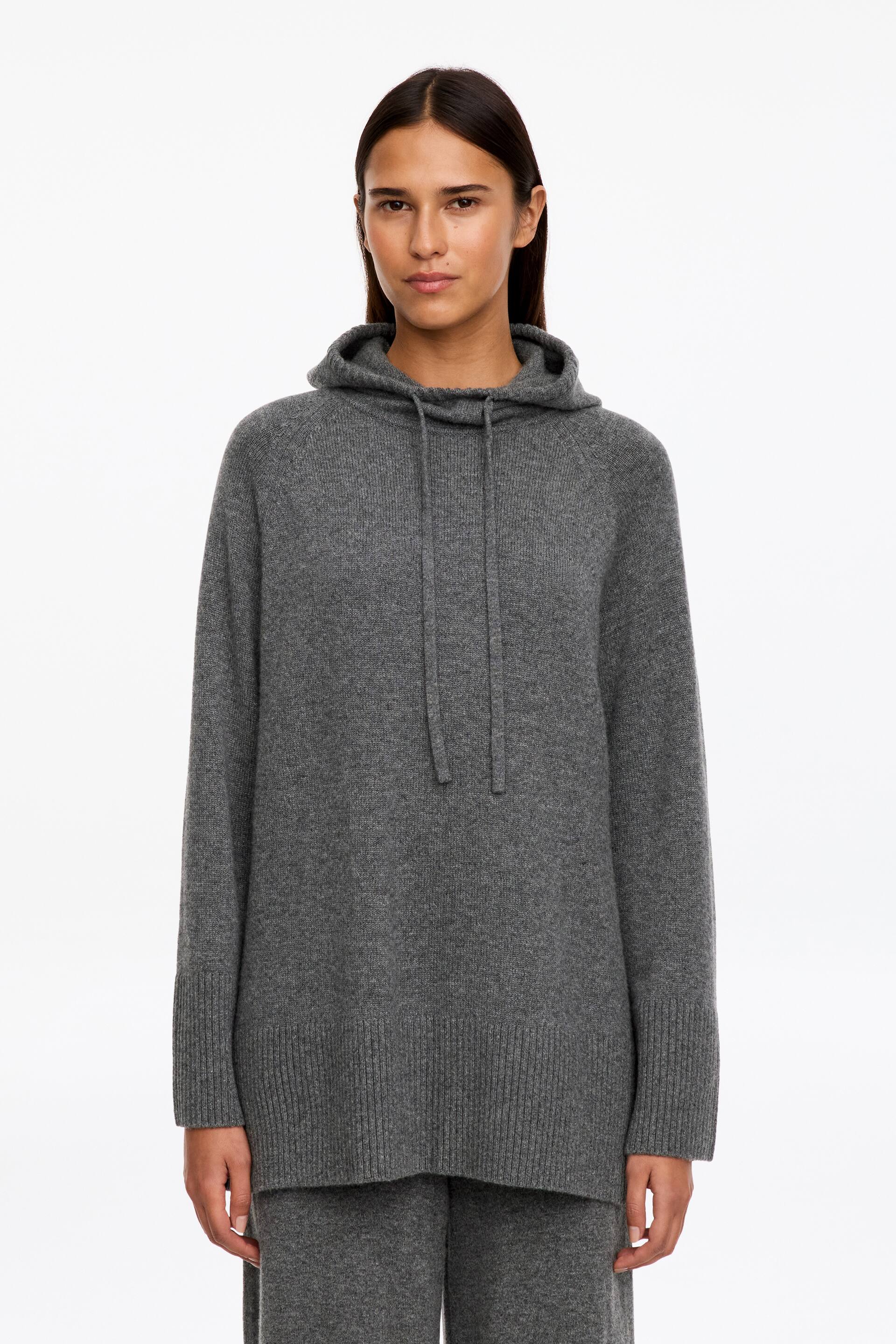 Vis større bilde: En grå kashmirhettegenser med lang silhuett bæres av modellen, og har justerbar hette med snøring, raglanermer og brede ribbestrikkede kanter ved mansjettene og nederkanten. Hettegenseren styles med matchende grå strikket bukse.