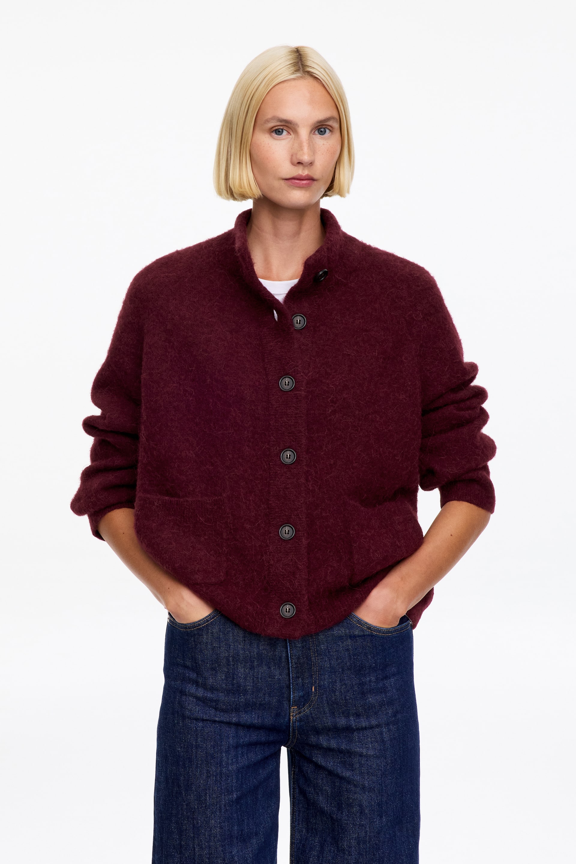 Visualizza immagine più grande: Un cardigan in maglia burgundy dalla vestibilità morbida e ampia, con una texture soffice e pelosa, indossato dalla modella sopra una t-shirt bianca e jeans blu scuro. Il cardigan presenta un colletto alto e una chiusura frontale con bottoni, mentre la modella ha le mani appoggiate nelle tasche dei jeans.