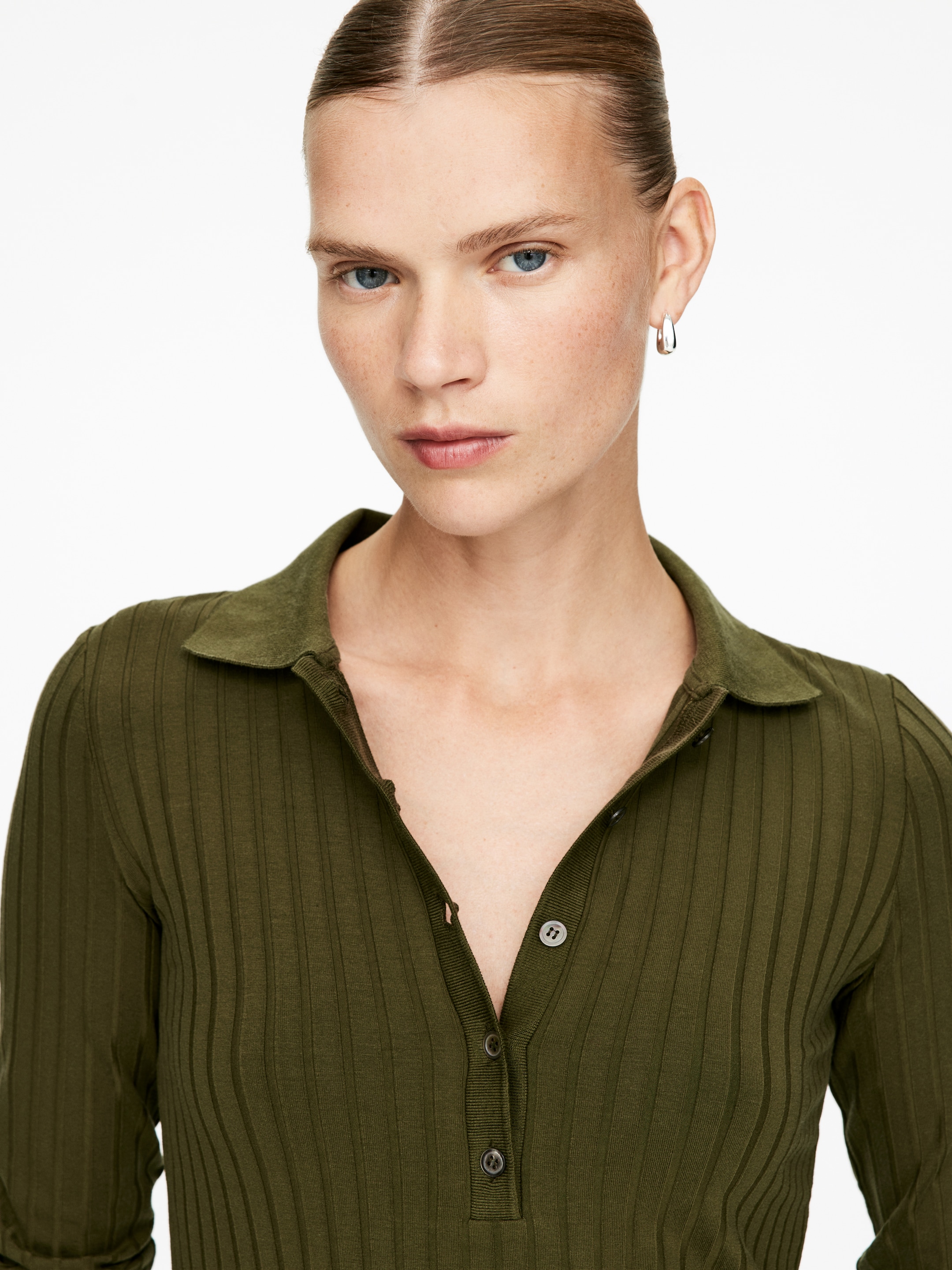 Größeres Bild ansehen: Ein olivgrünes langärmeliges Poloshirt wird getragen, das ein vertikal geripptes Strickmuster, einen klassischen Polokragen und eine Drei-Knopf-Leiste mit dunklen Knöpfen aufweist.
