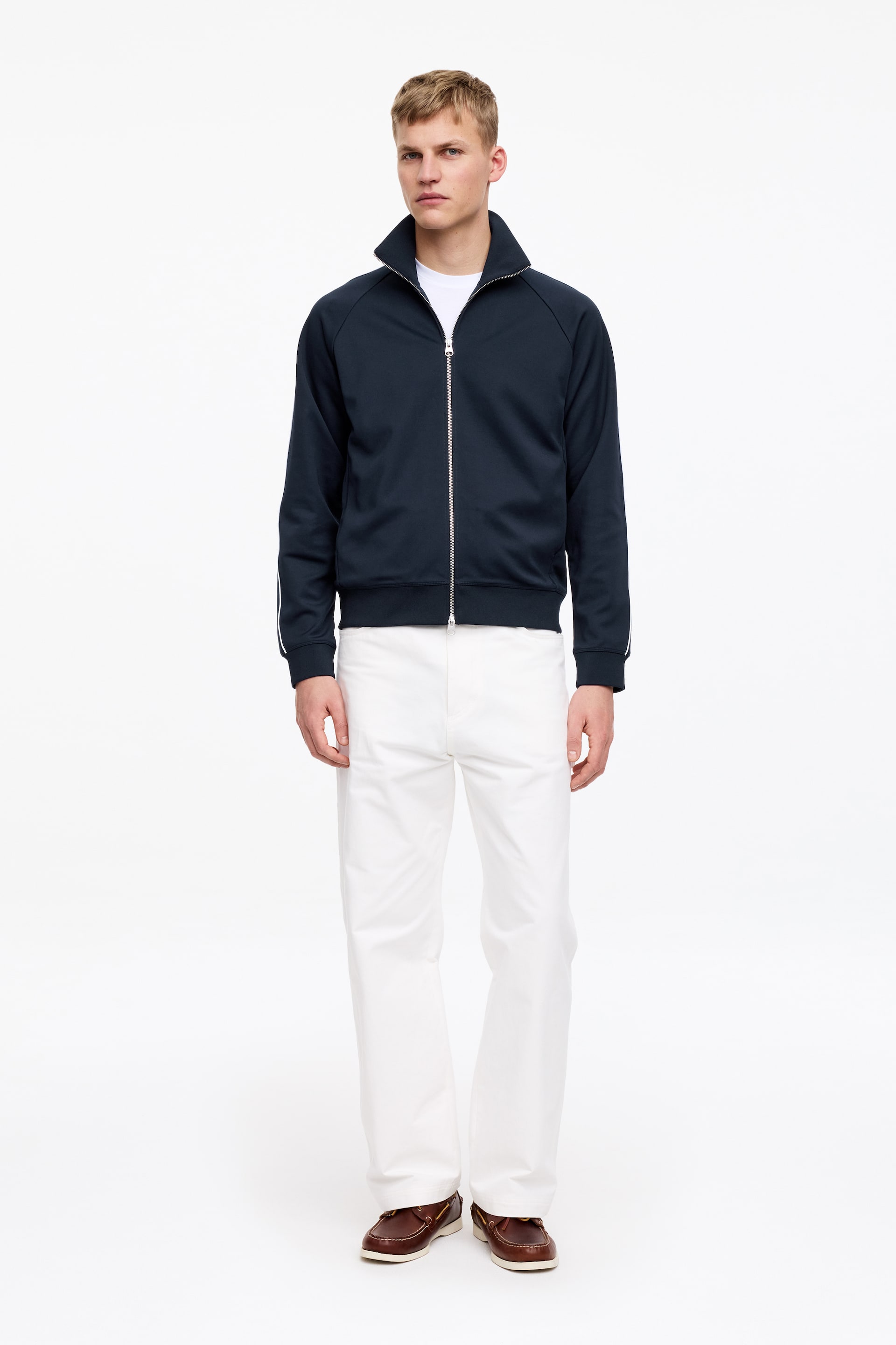 Agrandir l'image: Le mannequin porte une veste de survêtement bleu foncé, associée à un t-shirt blanc porté en dessous. La veste présente un passepoil blanc le long des manches, une fermeture éclair métallique argentée à double curseur sur le devant, des poches en biais et des finitions côtelées au niveau du col, des poignets et de l'ourlet.