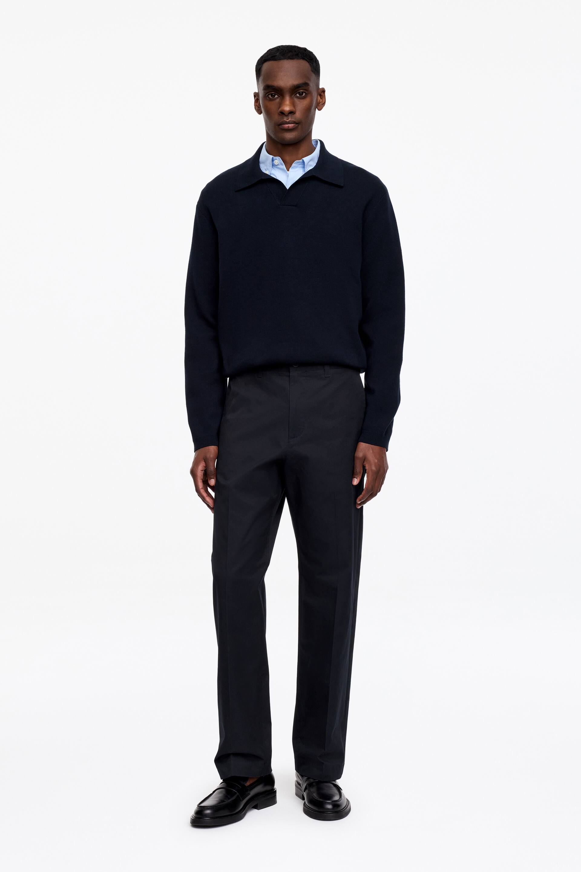Grotere afbeelding bekijken: Een donkerblauwe, gebreide polo met lange mouwen wordt frontaal gepresenteerd, met een open V-hals sluiting en gelaagd over een lichtblauw overhemd met kraag. Donkere, rechte broek met vouwen aan de voorkant maken de outfit compleet, samen met zwarte loafers met dikke zolen.