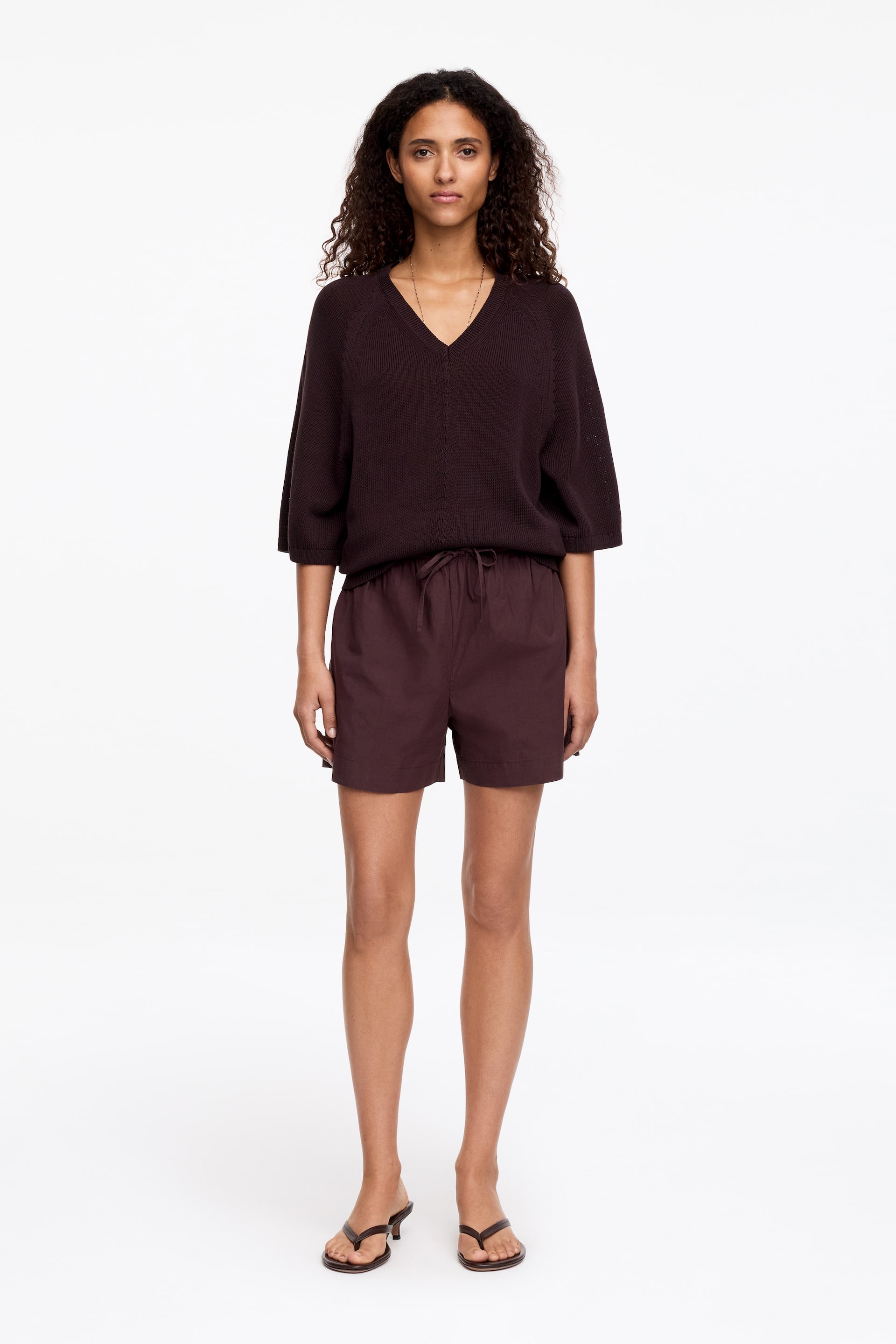 Agrandir l'image: Un pull marron foncé à col en V, doté de manches raglan trois-quarts et d'un col en V côtelé. Il est porté avec un short marron foncé assorti, qui présente une taille à cordon de serrage et une coupe décontractée, reposant au-dessus du genou.
