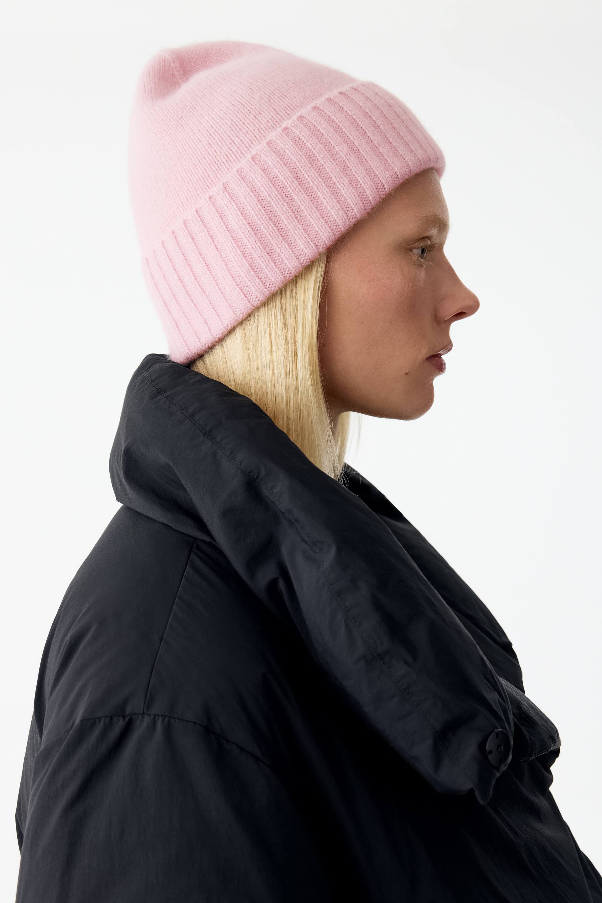 Grotere afbeelding bekijken: Een lichtroze gebreide beanie met een brede, omgeslagen ribboord wordt gedragen door het model, dat in zijaanzicht is afgebeeld. De beanie is gestyled in combinatie met een zwarte gewatteerde jas met hoge kraag.