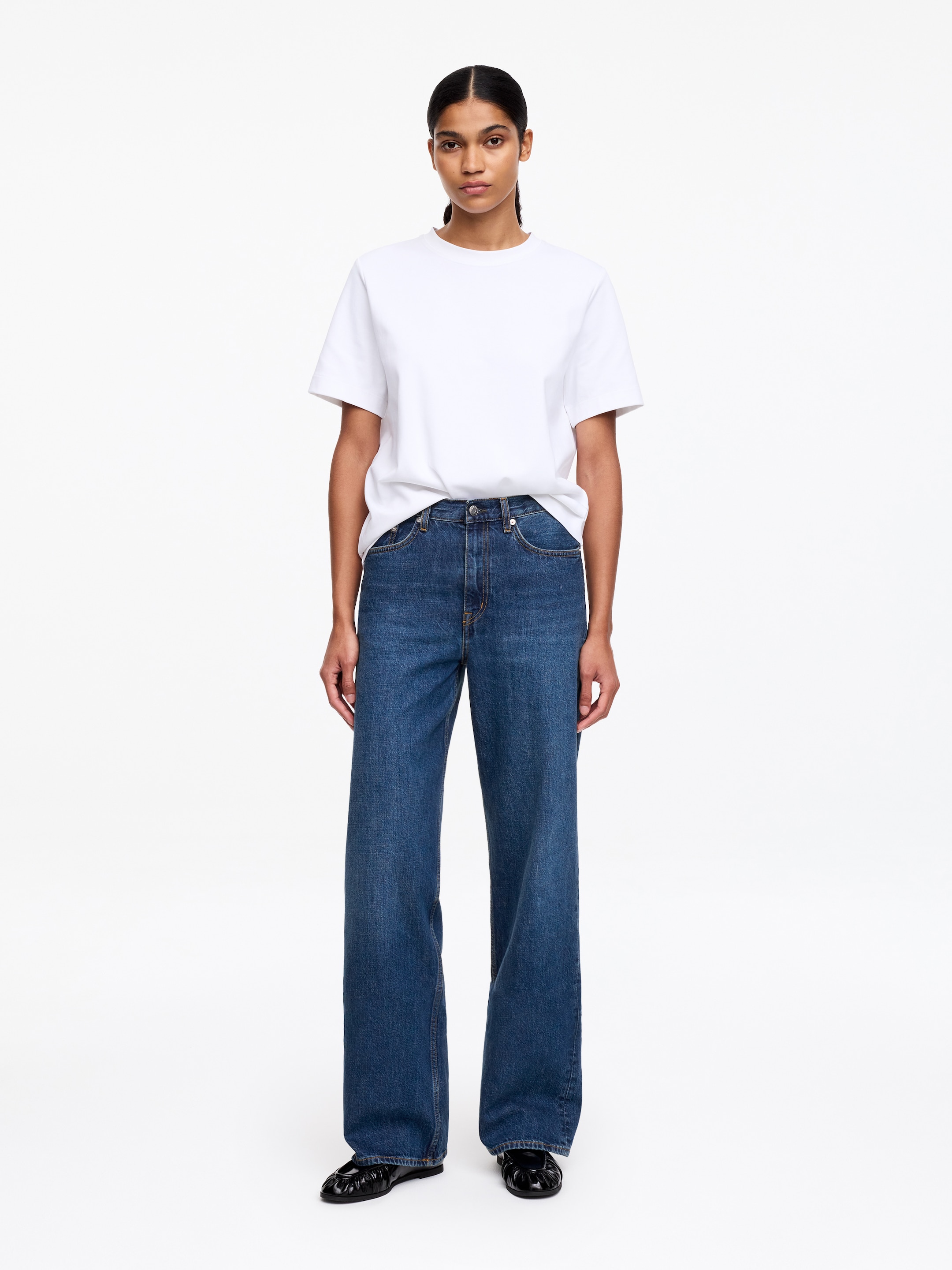 Ver imagem maior: Está a ser usada uma t-shirt branca de manga curta com gola redonda, parcialmente colocada dentro de umas calças de ganga azuis de lavagem média, de cintura alta e perna larga. Sapatos rasos pretos com detalhe franzido no peito do pé completam o conjunto.