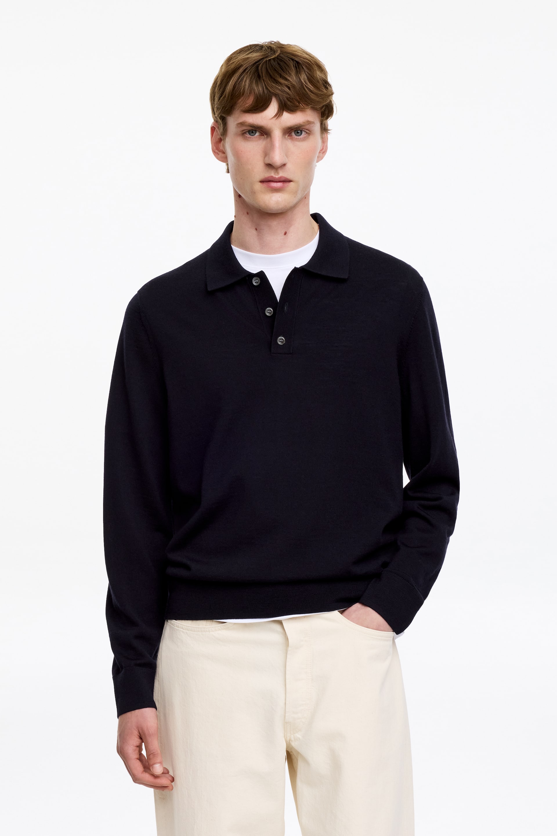 View larger image: A navy blue long-sleeved polo jumper featuring a polo collar and a three-button placket with dark buttons. It displays ribbed cuffs and a ribbed hem, and is worn layered over a white crew-neck t-shirt.