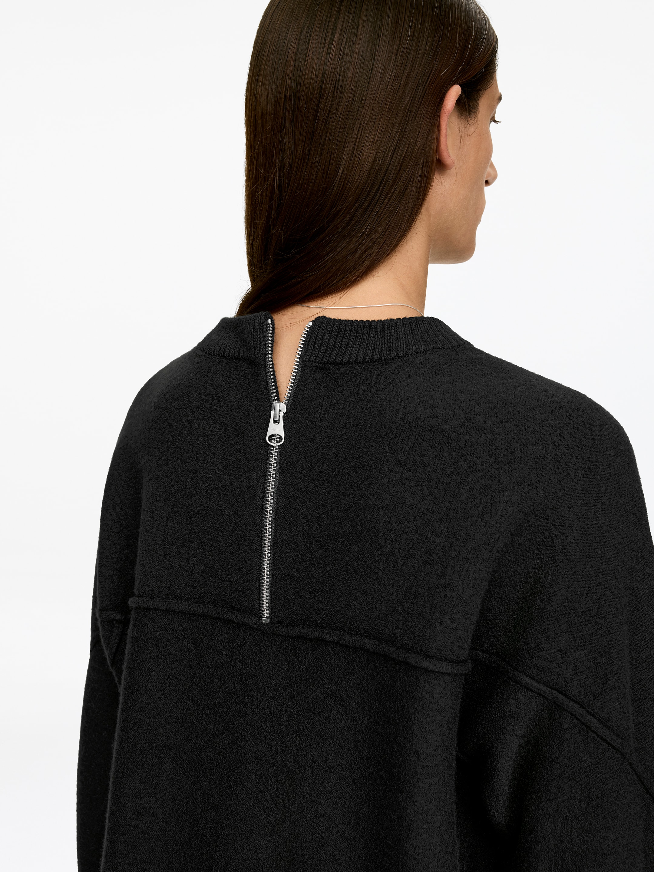 Visualizza immagine più grande: Un mini abito nero a vestibilità ampia realizzato in lana cotta, caratterizzato da maniche lunghe e una mezza zip sul retro, è indossato dalla modella con la zip parzialmente aperta.