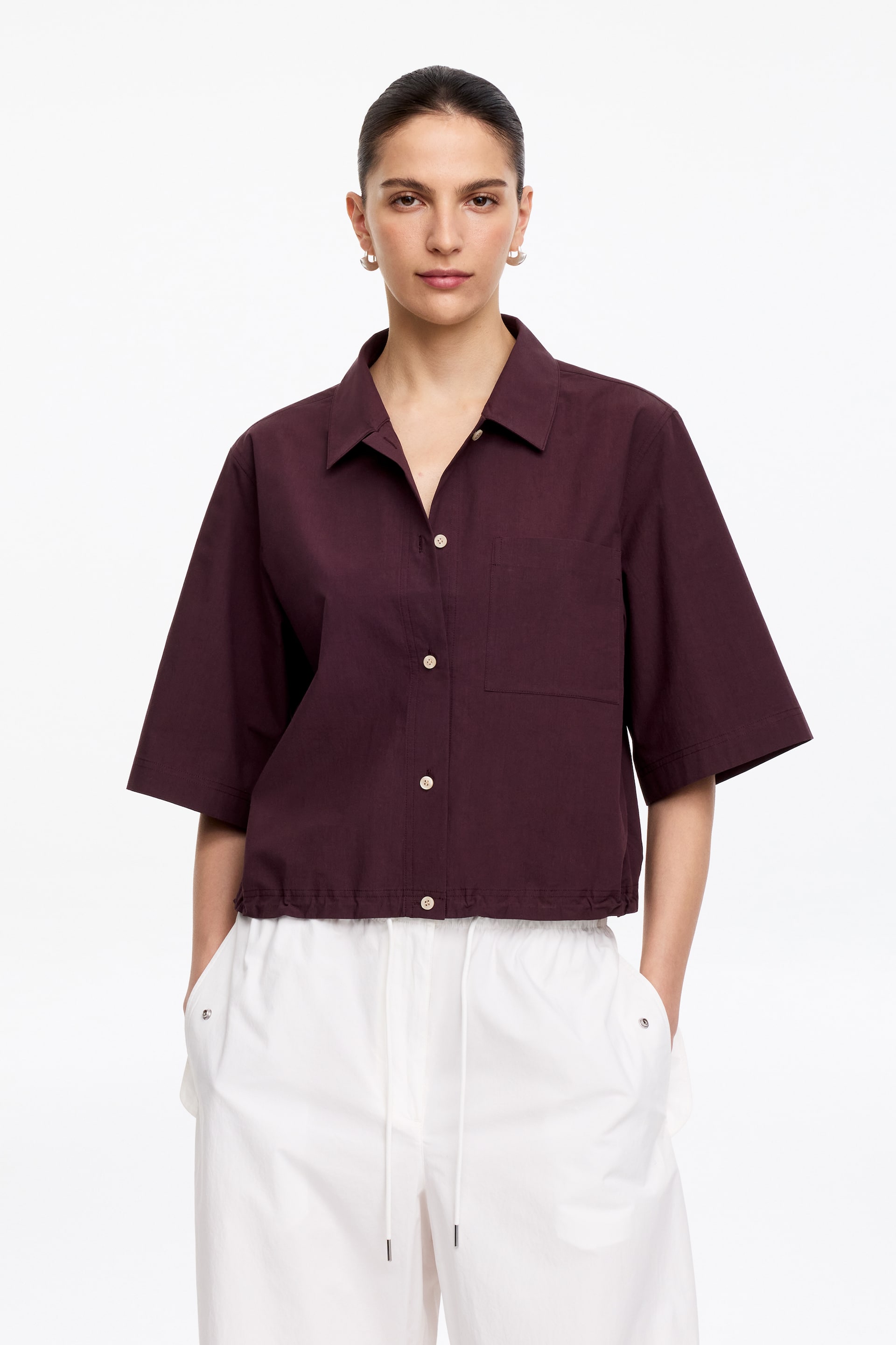 Agrandir l'image: La chemise à manches courtes rouge foncé présente un col évasé, une fermeture à boutons sur le devant avec des boutons de couleur claire, une poche plaquée unique sur la poitrine gauche, des manches amples et un ourlet élastiqué et raccourci.