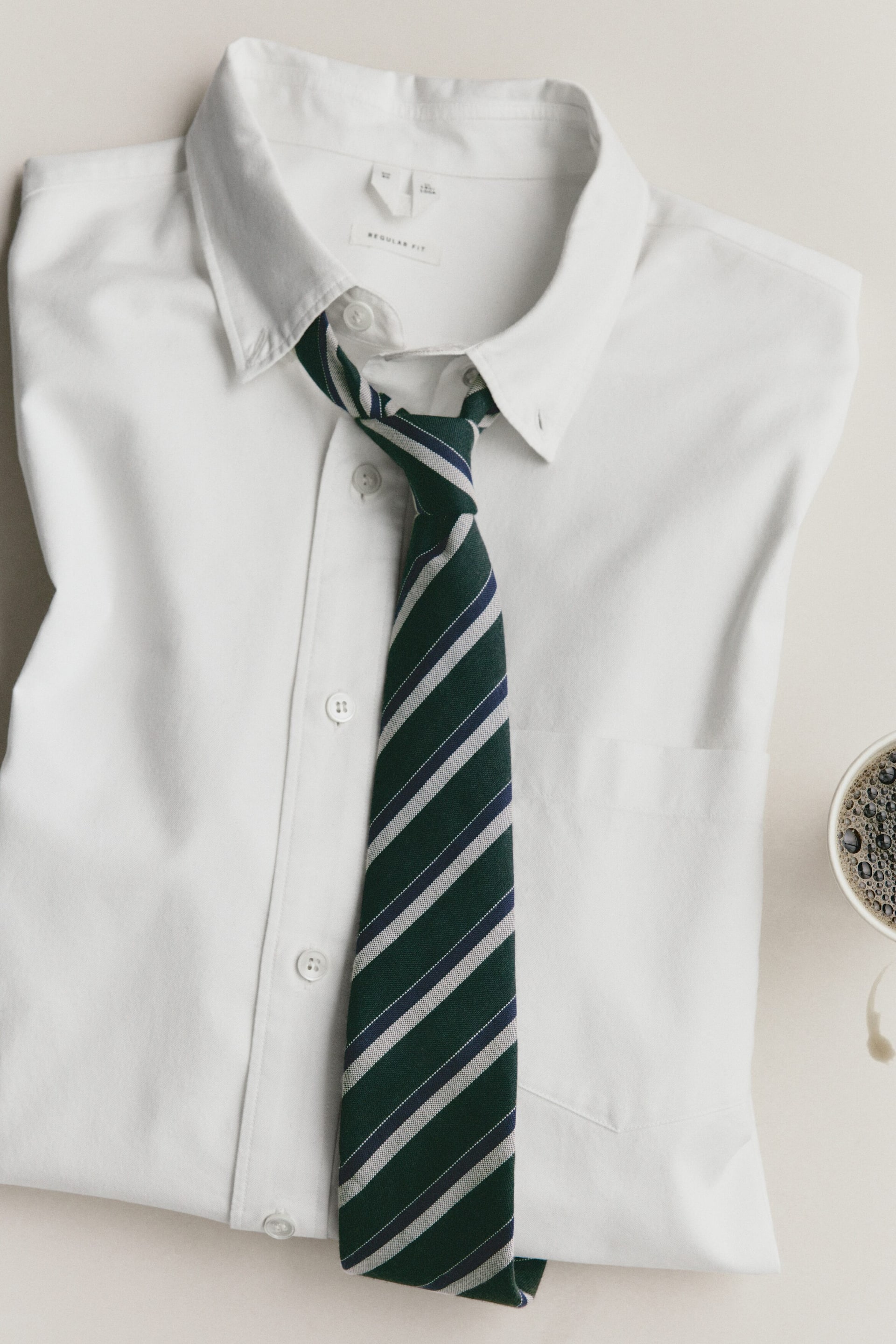 Ver imagem maior: Uma camisa social branca de manga comprida dobrada está estendida, exibindo o design frontal com gola button-down, carcela de botões e bolso no peito. Uma gravata verde-escura com riscas diagonais em azul-marinho, cinzento e branco está atada de forma solta sob a gola.