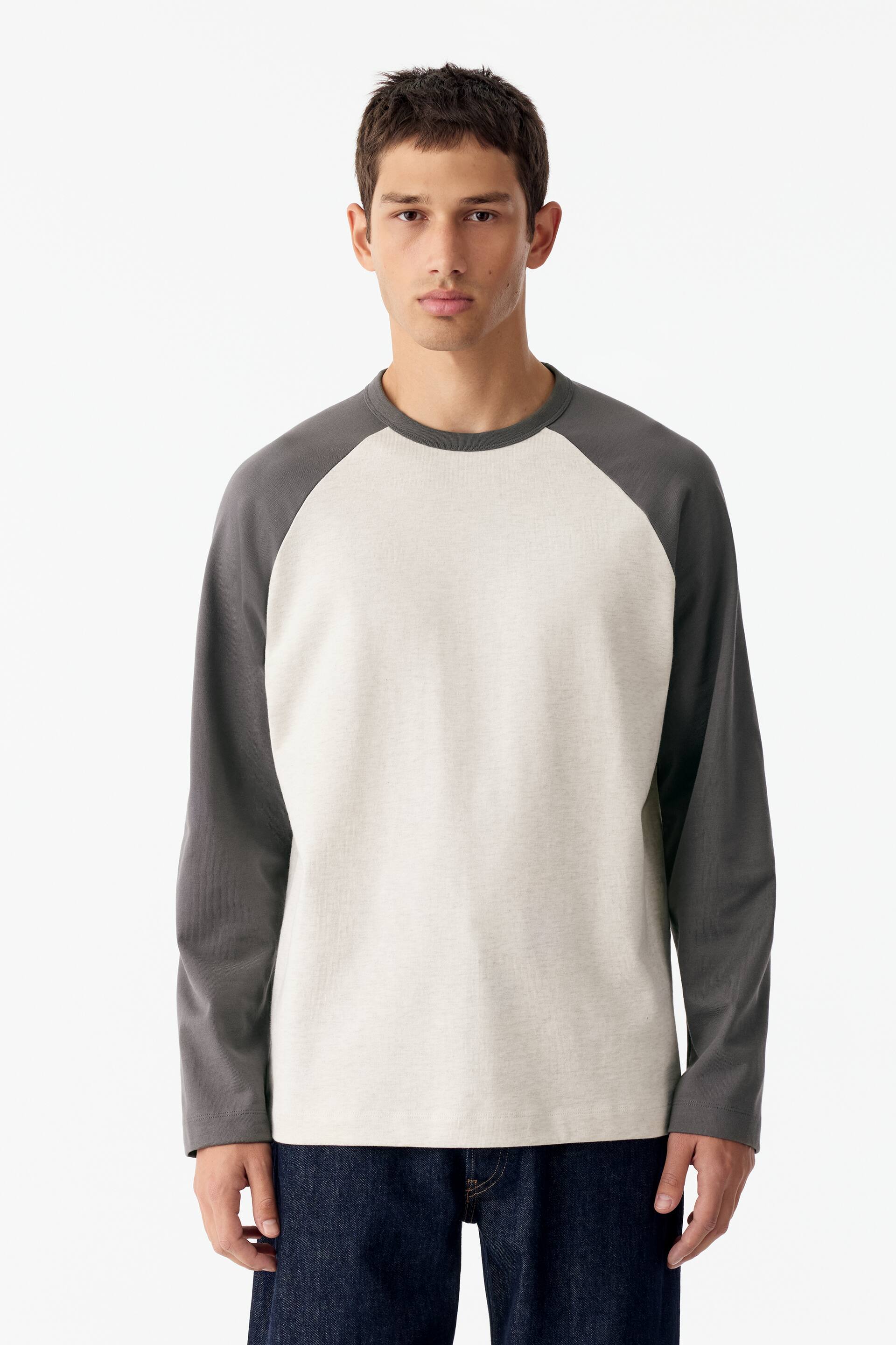 Vezi imaginea mai mare: Un tricou de baseball cu corp de culoarea crem și mâneci raglan lungi de culoare gri închis este purtat de model. Tricoul are un guler rotund rib tricotat, asortat, de culoare gri închis, și este combinat cu blugi bleumarin închis.
