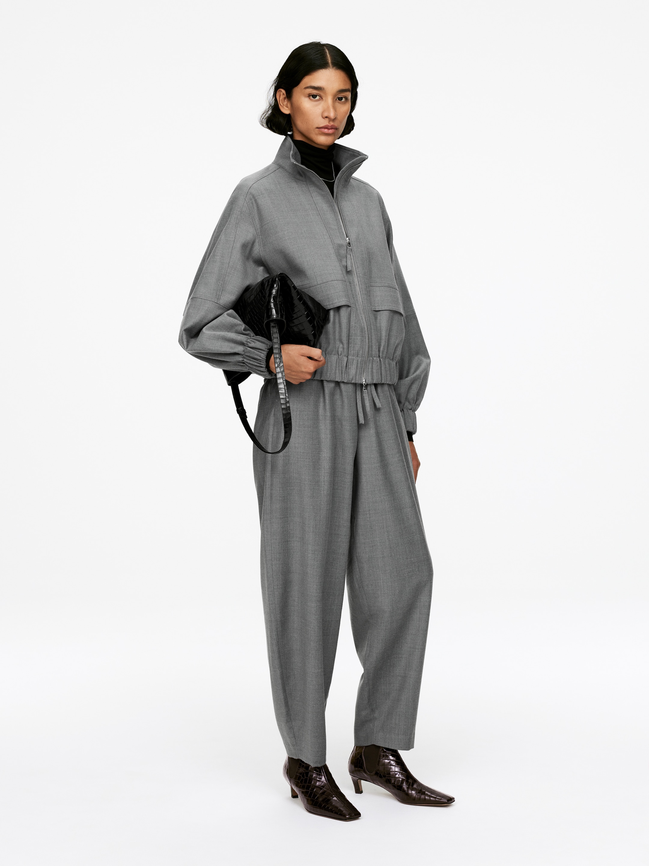 Visa större bild: En grå tvådelad outfit presenteras i en framåtvänd vy, bestående av en jacka med full dragkedja och hög ståkrage, voluminösa ärmar, horisontella bröstveck och en elastisk fåll, tillsammans med matchande avslappnade, vida byxor med dragsko i midjan. En svart polotröja är synlig under, och en svart texturerad kuvertväska samt mörkbruna texturerade ankelstövlar kompletterar ensemblet.