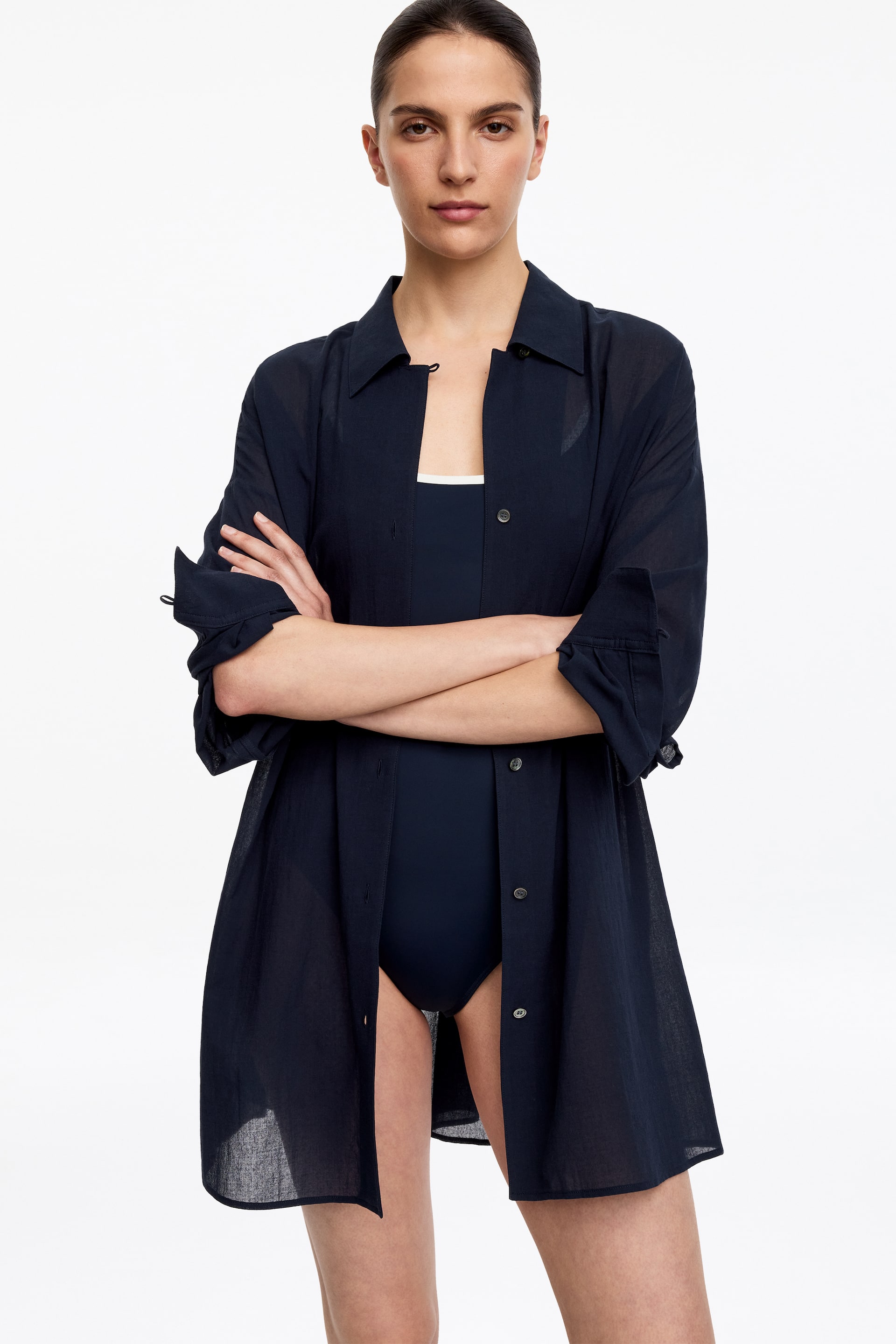 Agrandir l'image: Une robe-chemise transparente bleu marine est présentée de face, portée ouverte sur un maillot de bain foncé avec une bretelle blanche visible. Son col pointu, ses manches longues retroussées et fixées, ainsi que sa silhouette ample tombant jusqu'à mi-cuisse sont visibles.