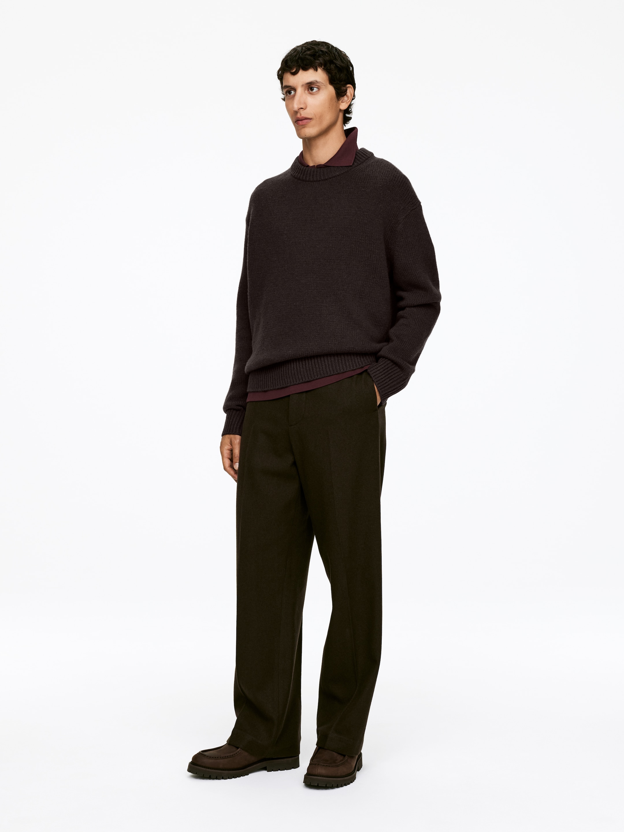 Größeres Bild anzeigen: Ein Model trägt einen dunkelbraunen Strickpullover mit Rundhalsausschnitt, entspannter Passform und langen Ärmeln, der über einem burgunderfarbenen Hemd mit Kragen geschichtet ist. Das Ensemble, vervollständigt mit dunkeloliven, weit geschnittenen Hosen und dunkelbraunen Loafern mit dicker Sohle, wird aus einem leichten Winkel präsentiert, um den entspannten Fall der Kleidungsstücke zu zeigen.