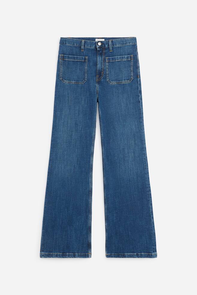A pair of high-rise blue denim jeans is shown from the front, featuring two large square patch pockets on the upper thighs and a classic button fastening with a zip fly. The jeans have a wide-leg silhouette with visible orange topstitching along the seams and hems. - 1